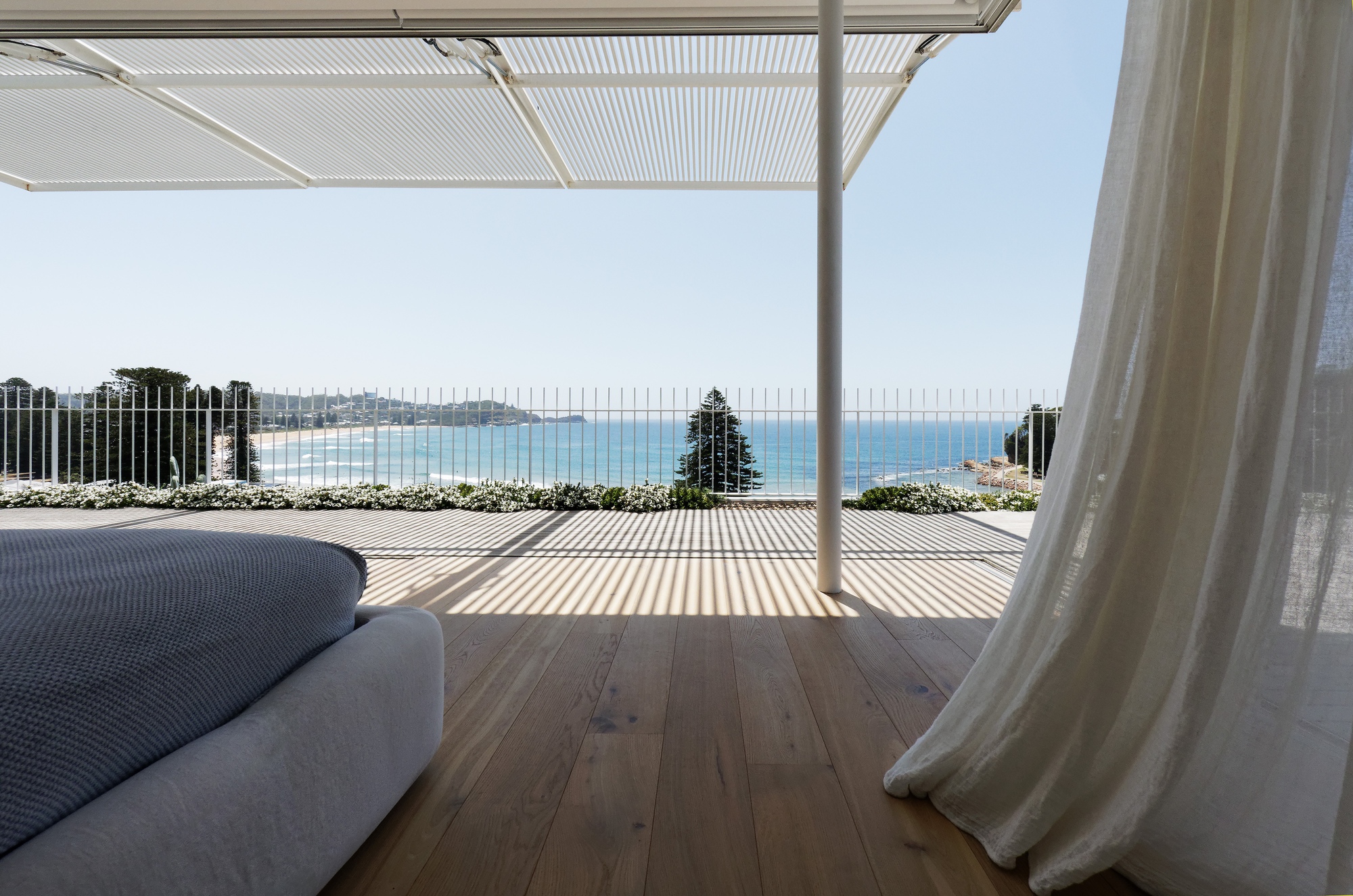 Covered terrace with timber decking and slatted canopy overlooking the ocean beyond a steel balustrade