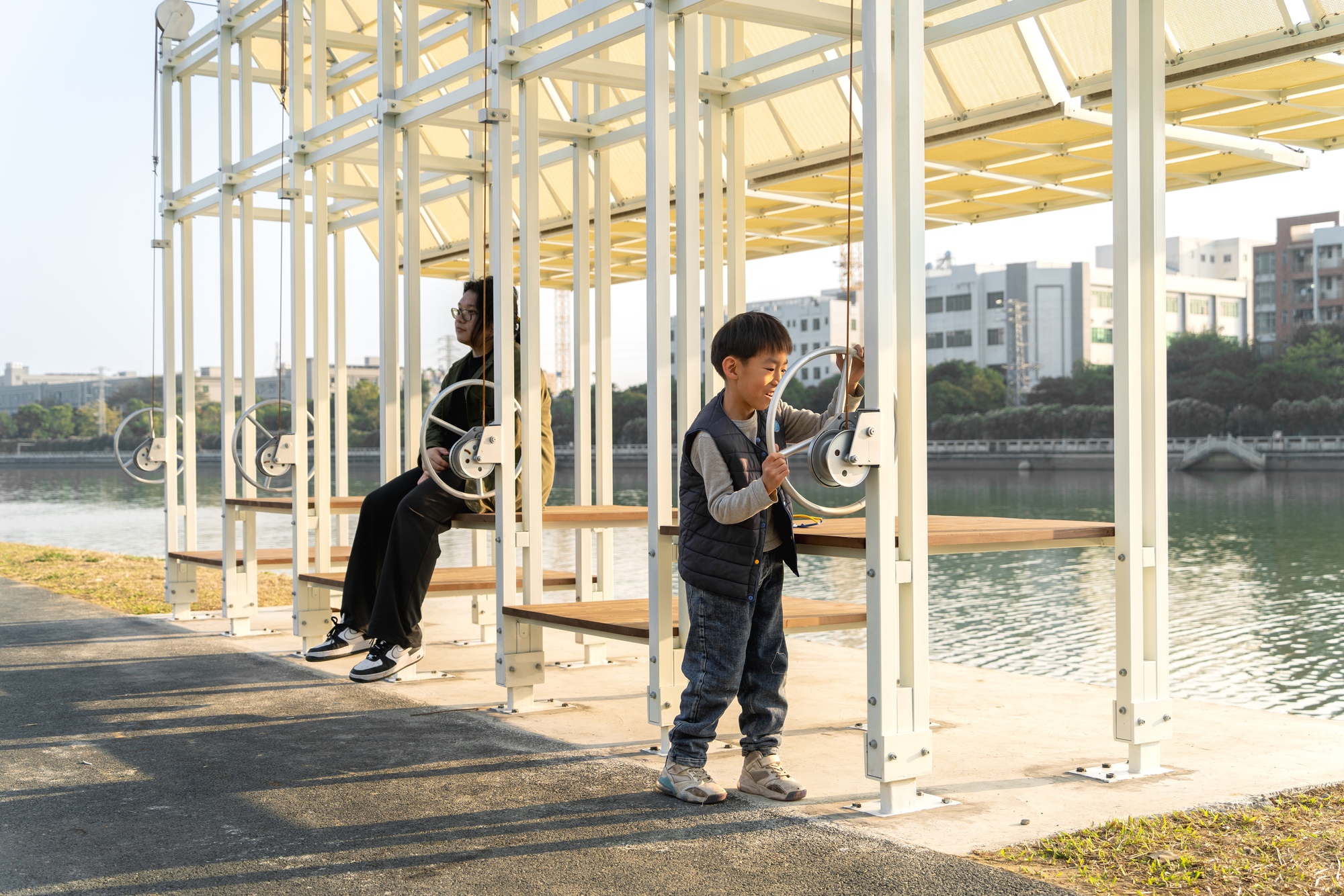 Children playing inside the white steel frame pavilion with river view beyond the columns