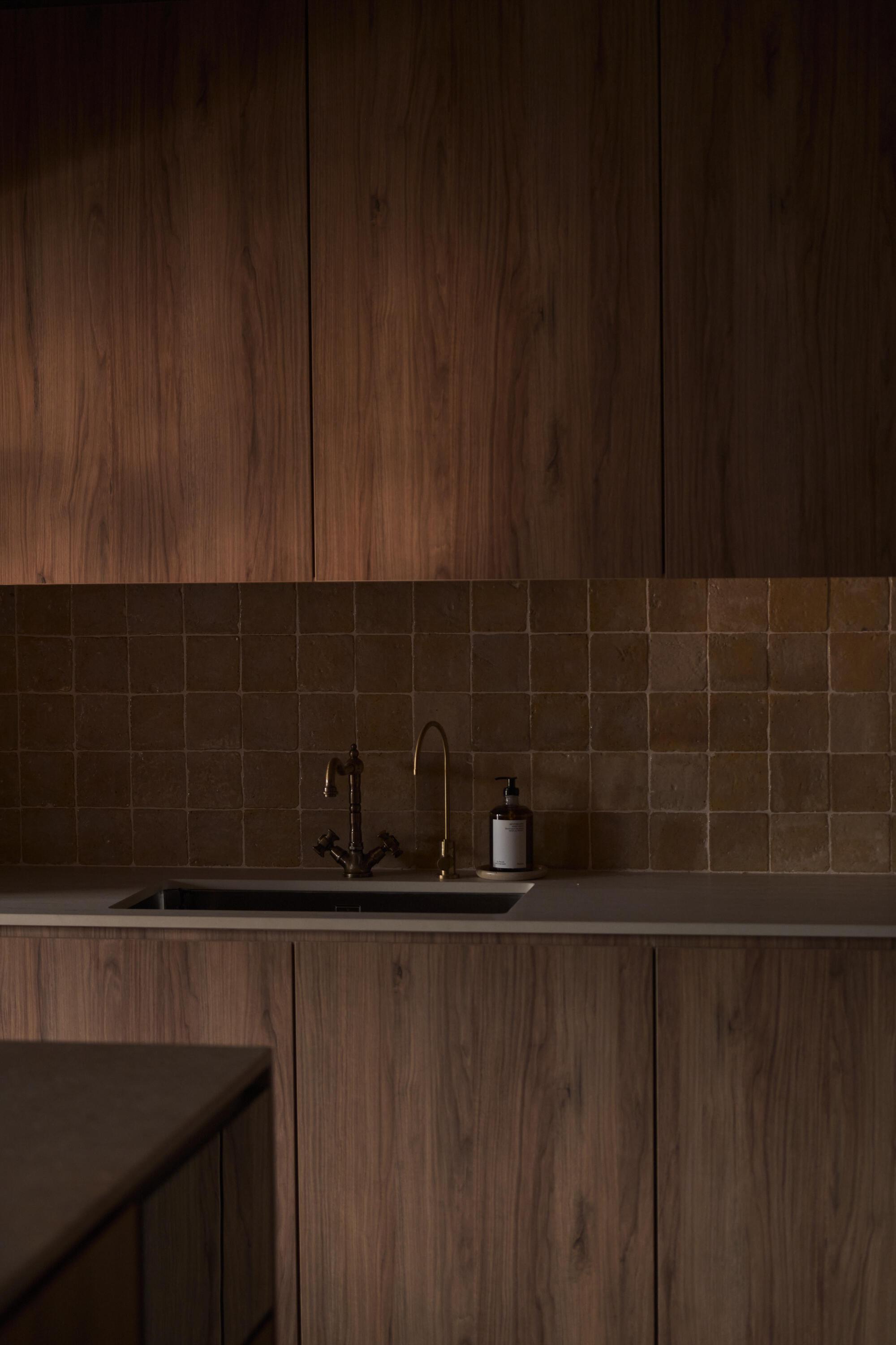 Kitchen counter with earthen tile backsplash below walnut veneer cabinetry in low light