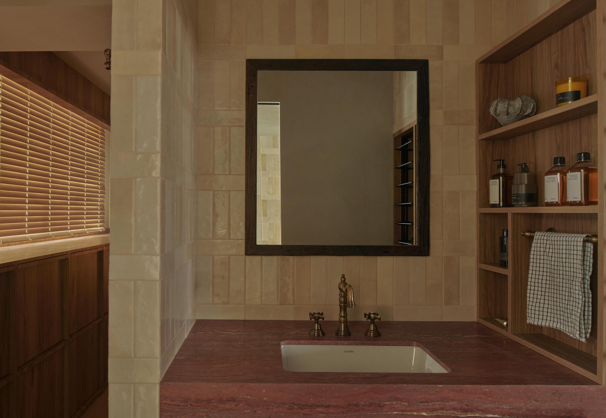 Bathroom vanity with red stone countertop, framed mirror and timber shelving beside a slatted timber blind