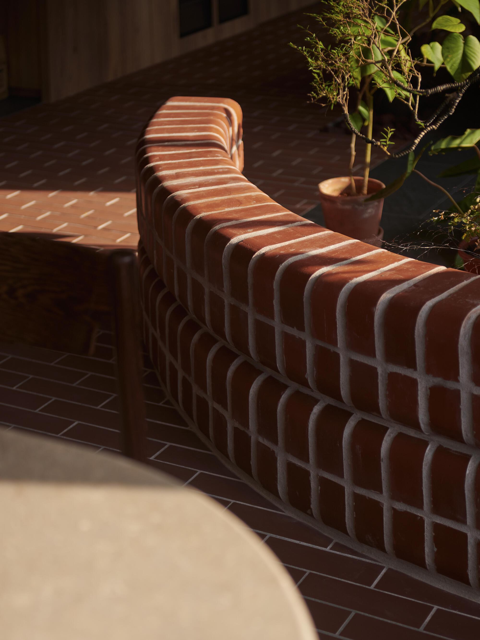 Curved terracotta brick planter wall with shadow patterns cast by overhead slats and potted tree
