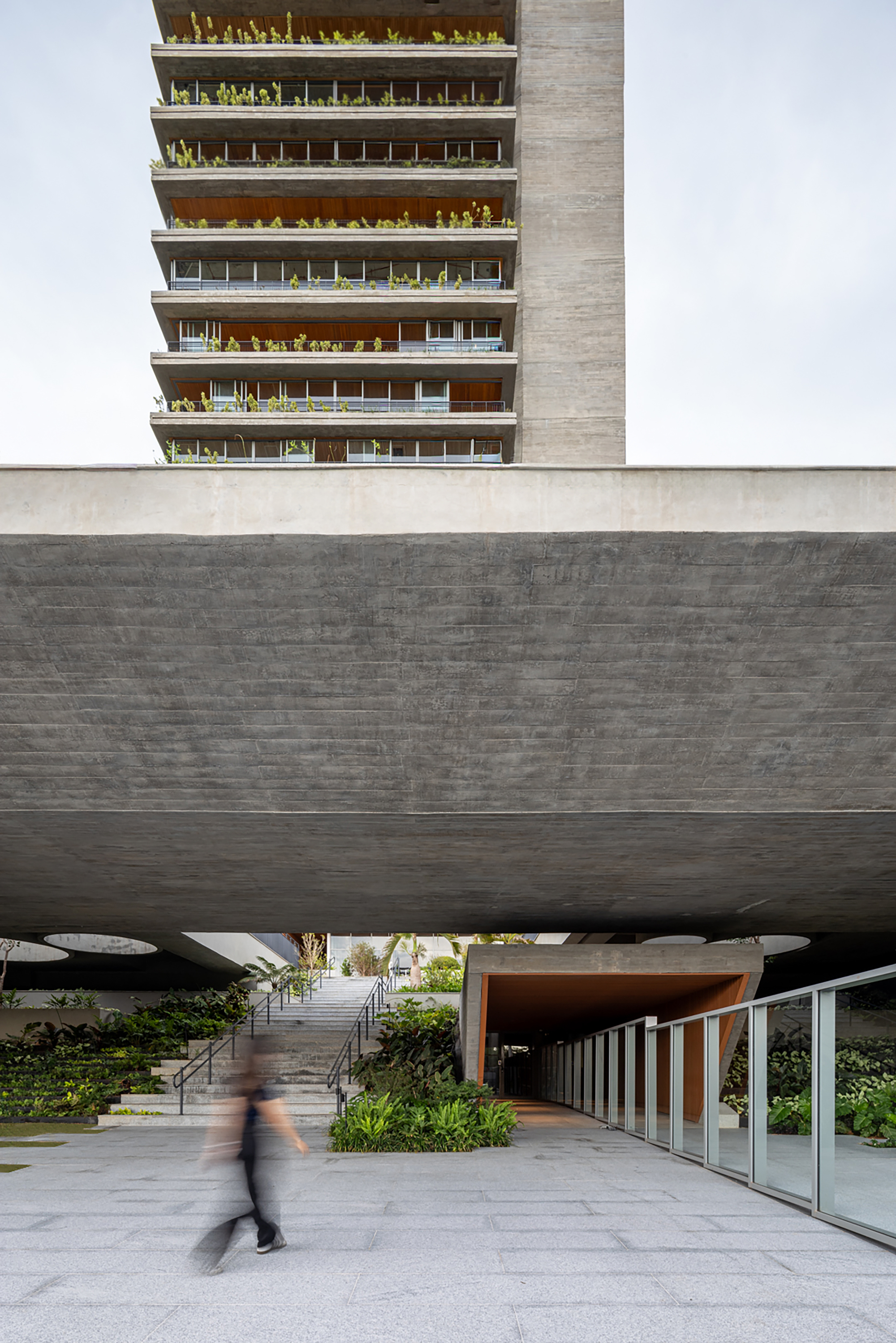 Cantilevered concrete podium deck framing the entrance stairway with a blurred figure crossing below