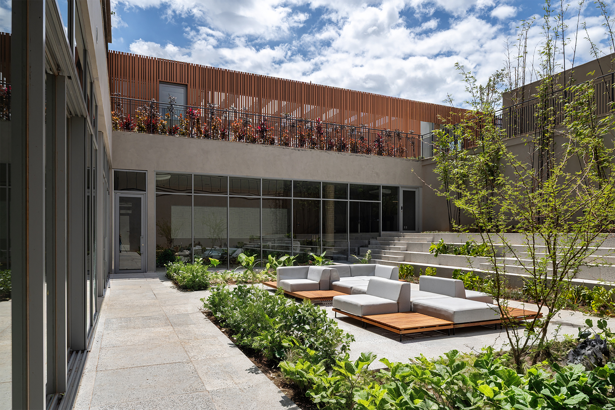 Courtyard terrace with timber and cushioned seating modules amid planted beds under blue sky