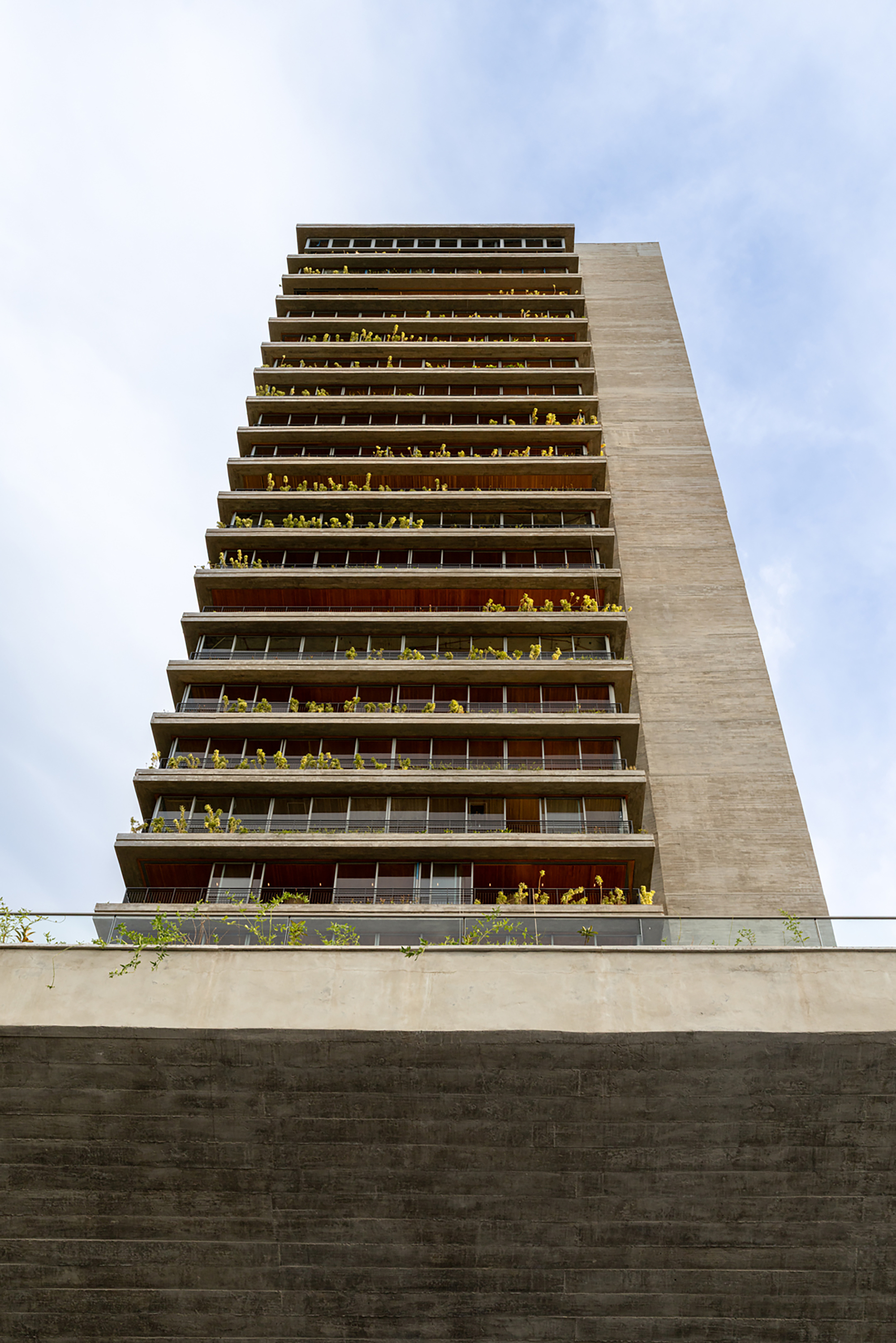 Upward view of a residential tower facade with planted balconies on each floor against a hazy sky