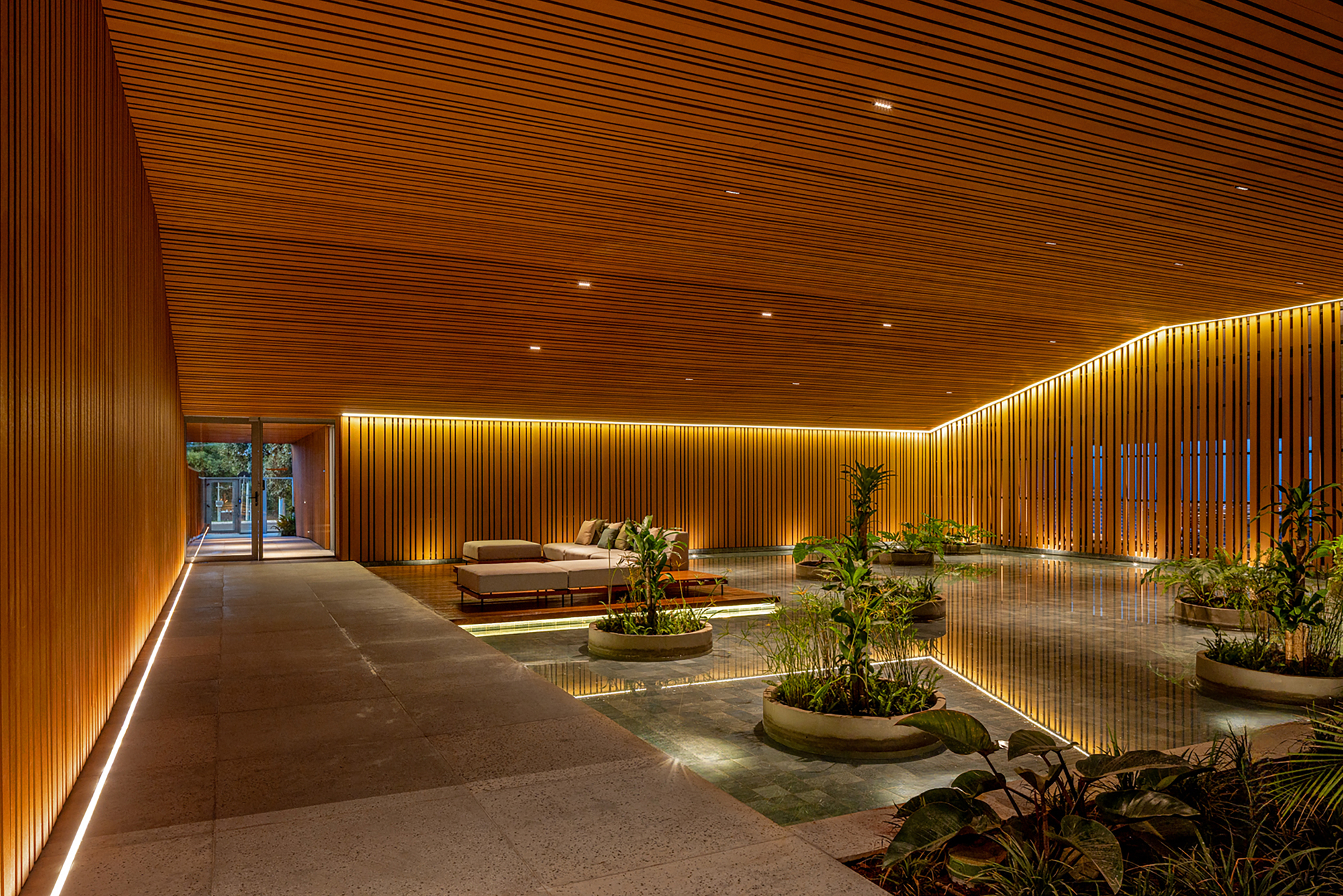 Interior lobby with linear wood ceiling, backlit vertical slat walls, and planted beds on polished stone flooring