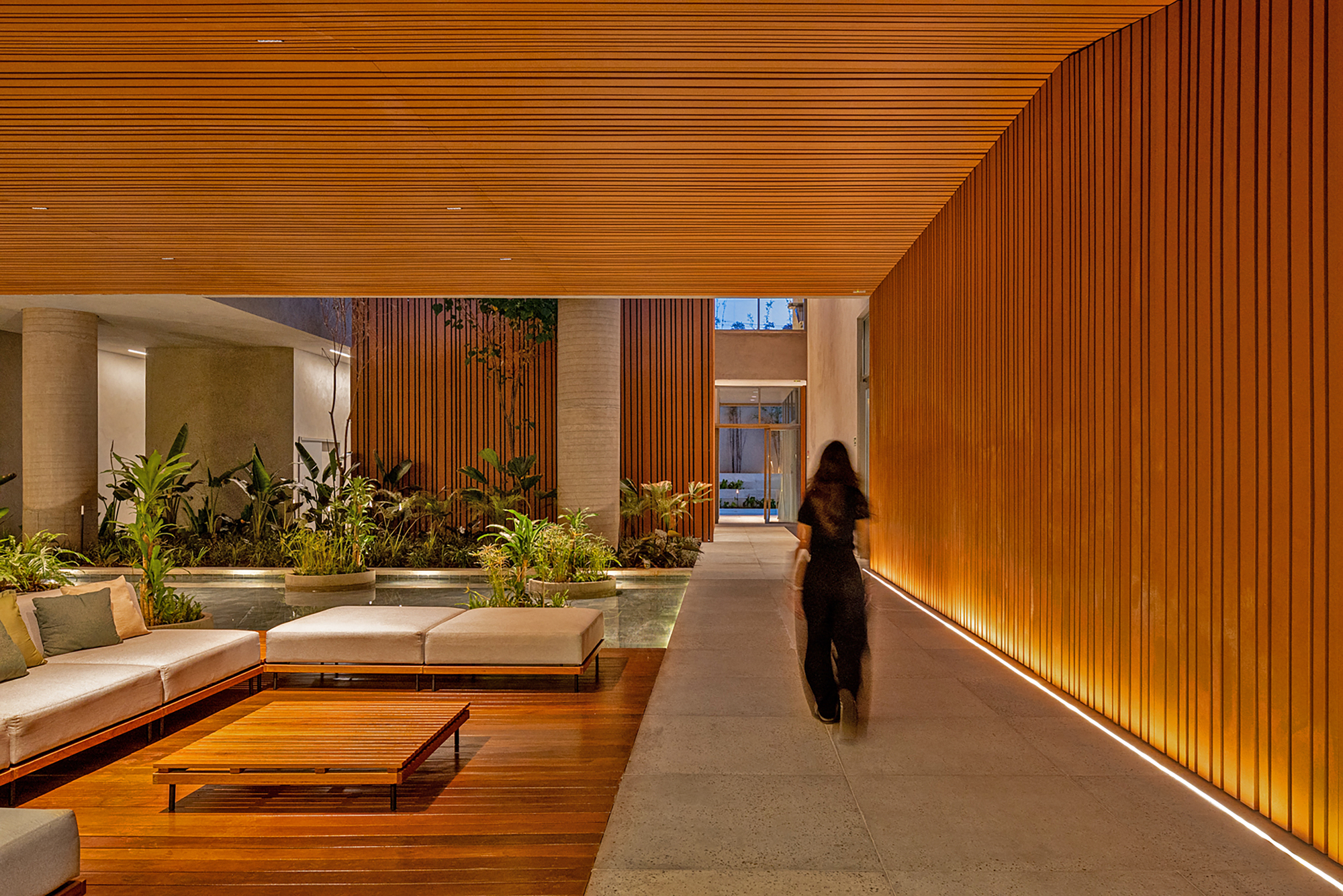 Circulation corridor with figure in motion passing illuminated wood slat wall and planted garden beds