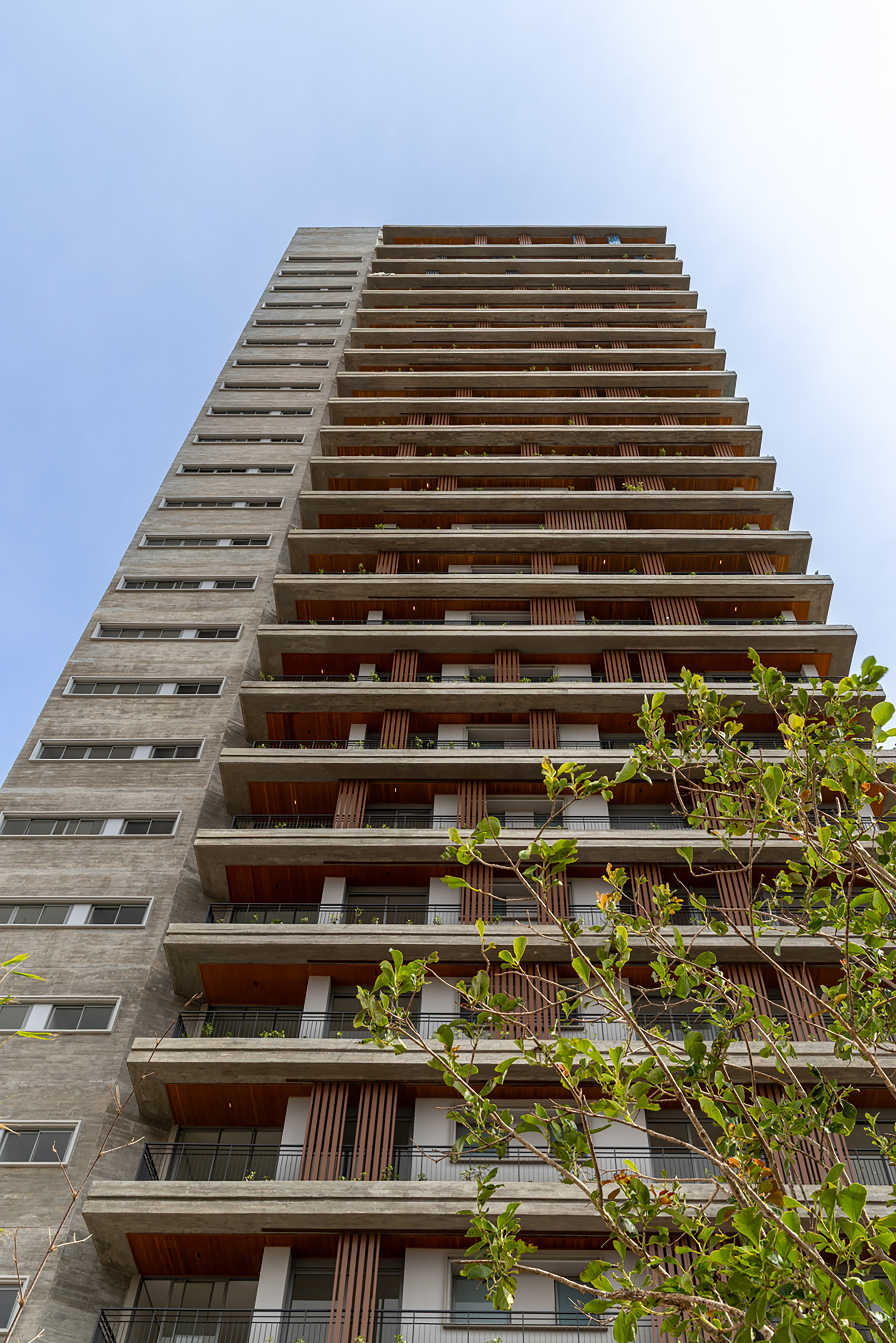 Upward view of residential tower facade with alternating concrete fins and timber-screened balconies against pale sky
