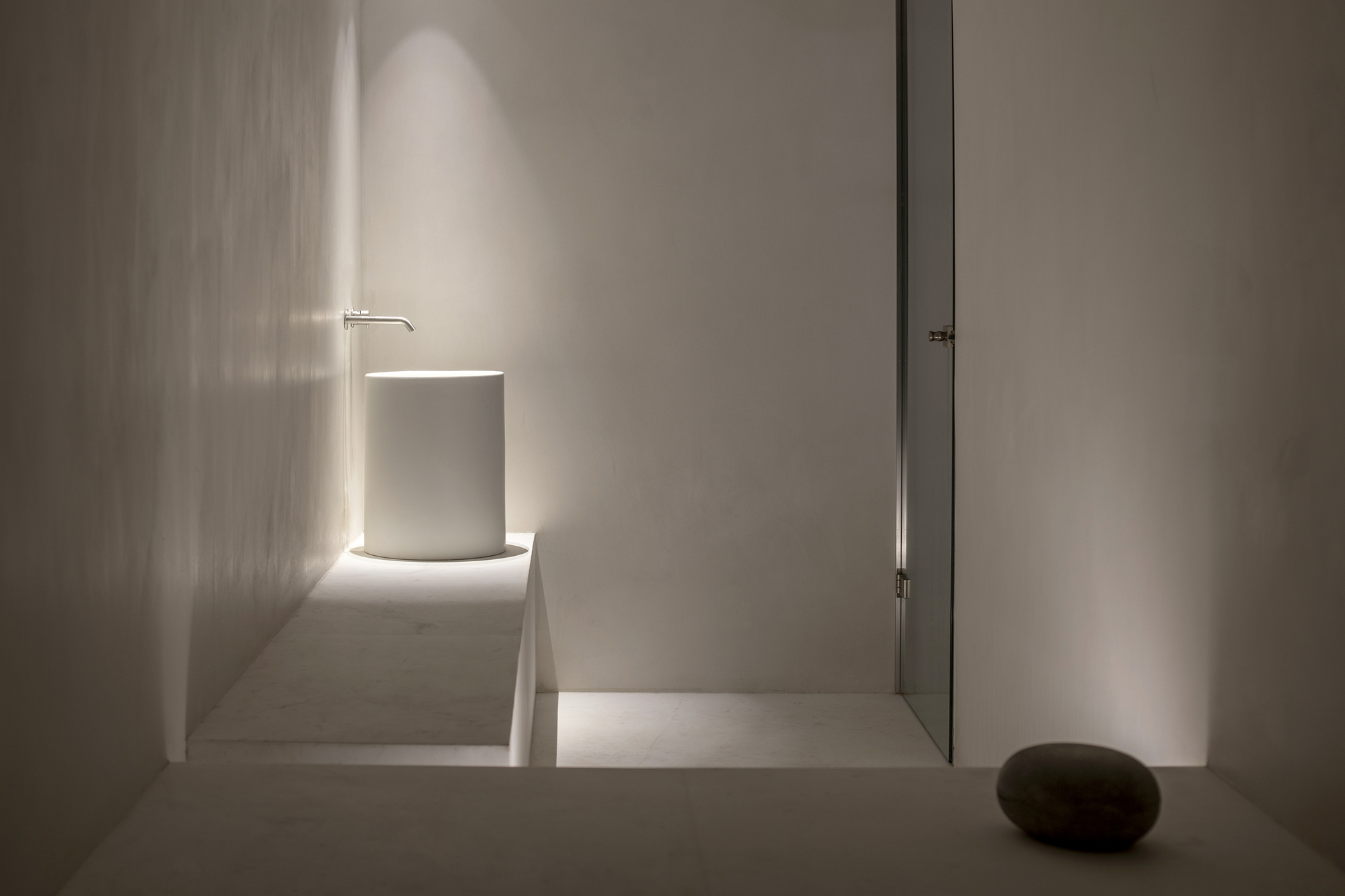 Minimalist corridor with backlit cylindrical volume and narrow slot of light along white plaster walls