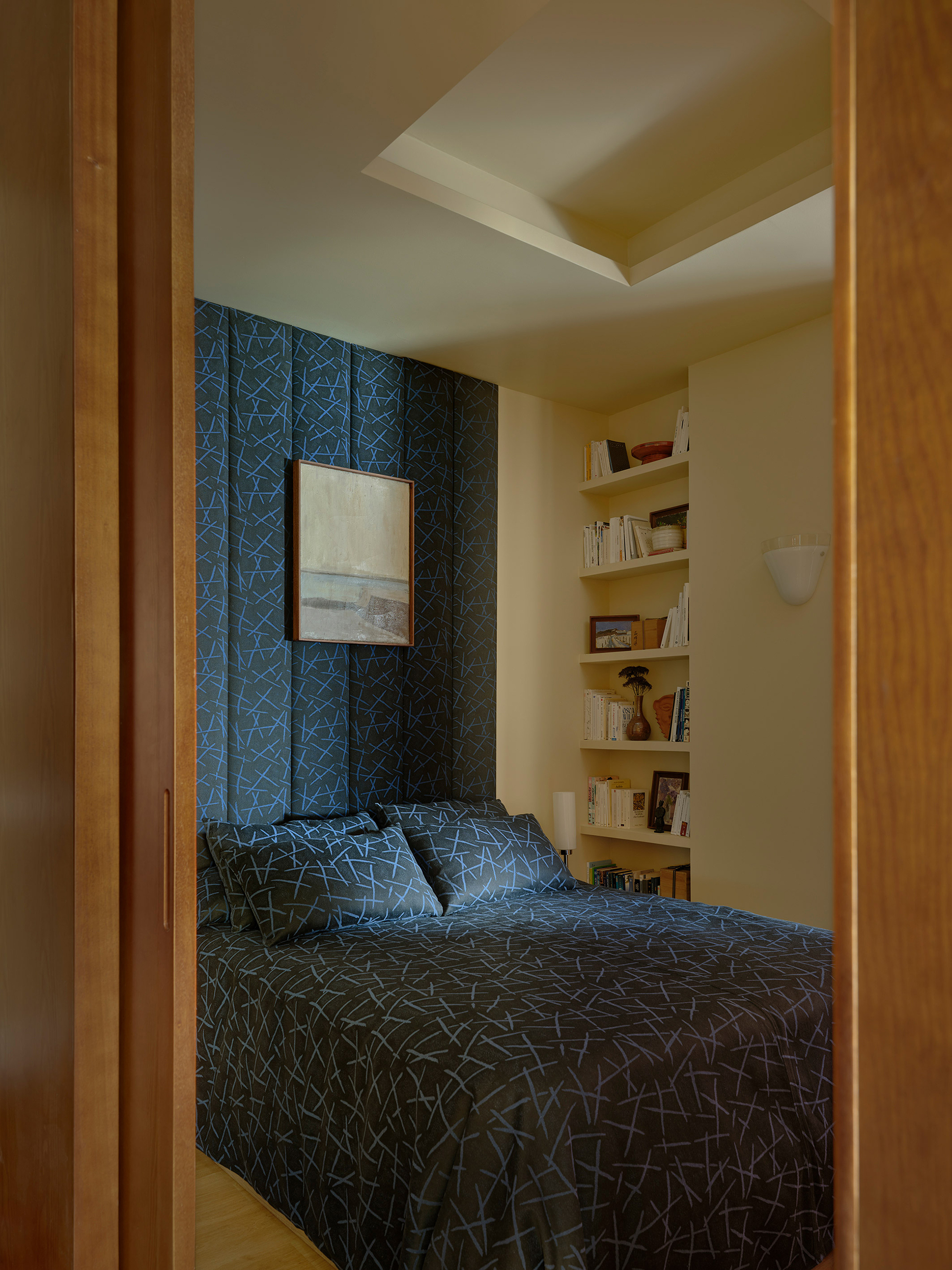 Bedroom alcove with patterned textile bedding and built-in shelving viewed through timber doorway