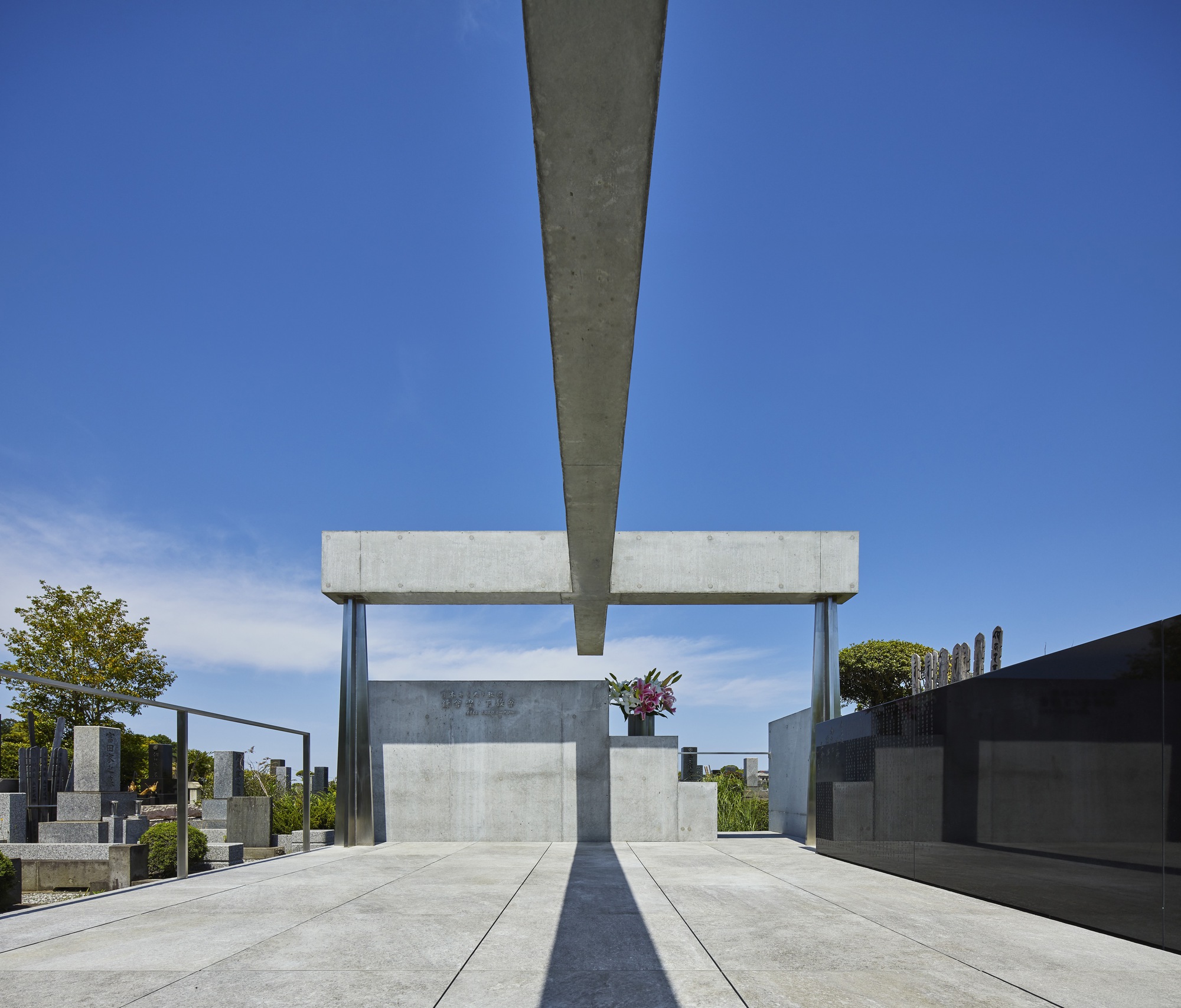 View along concrete plaza with central monument beneath cantilevered beam casting sharp shadows at midday