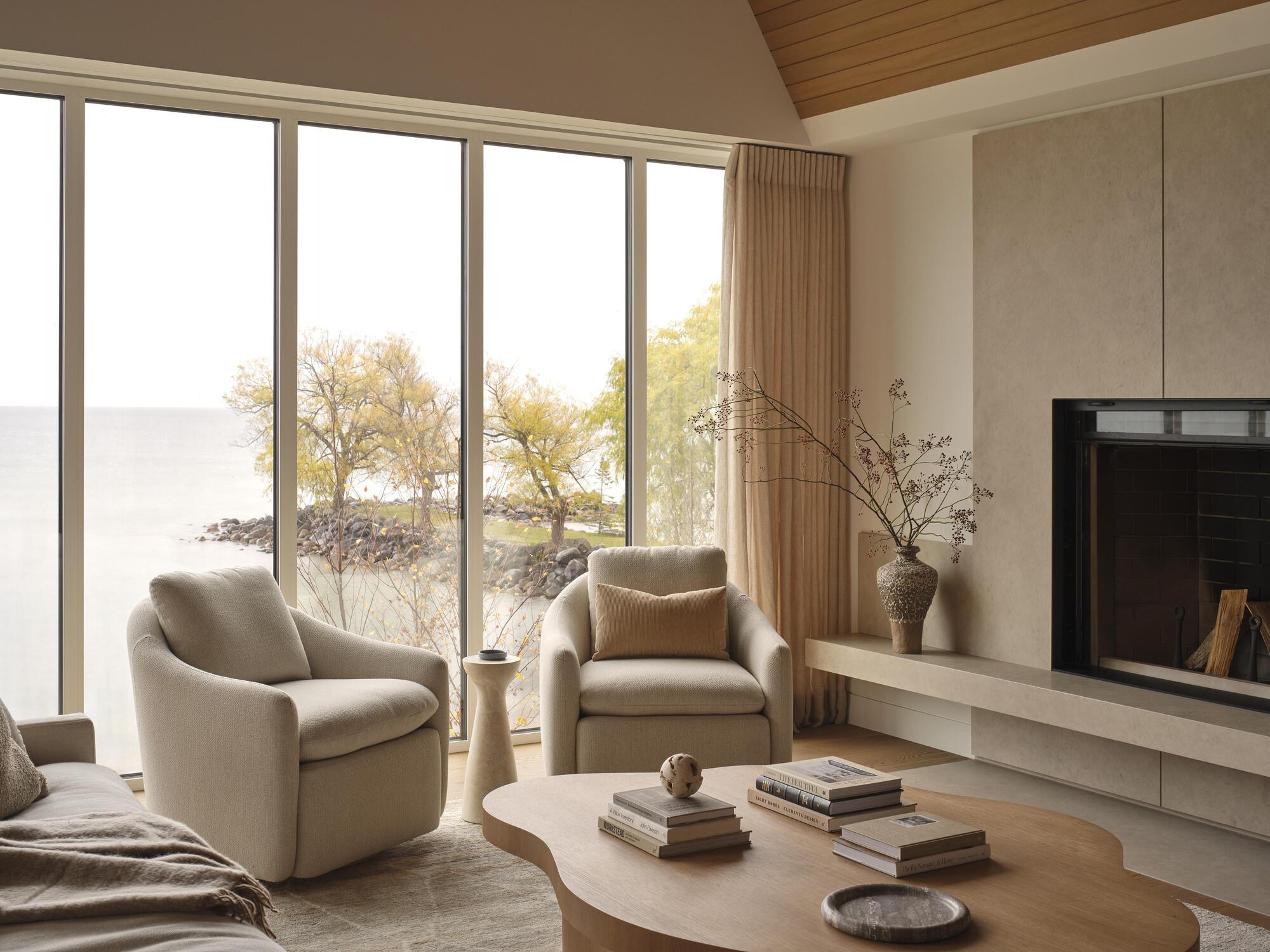 Seating area with fireplace hearth and floor-to-ceiling windows framing autumn trees along the shoreline