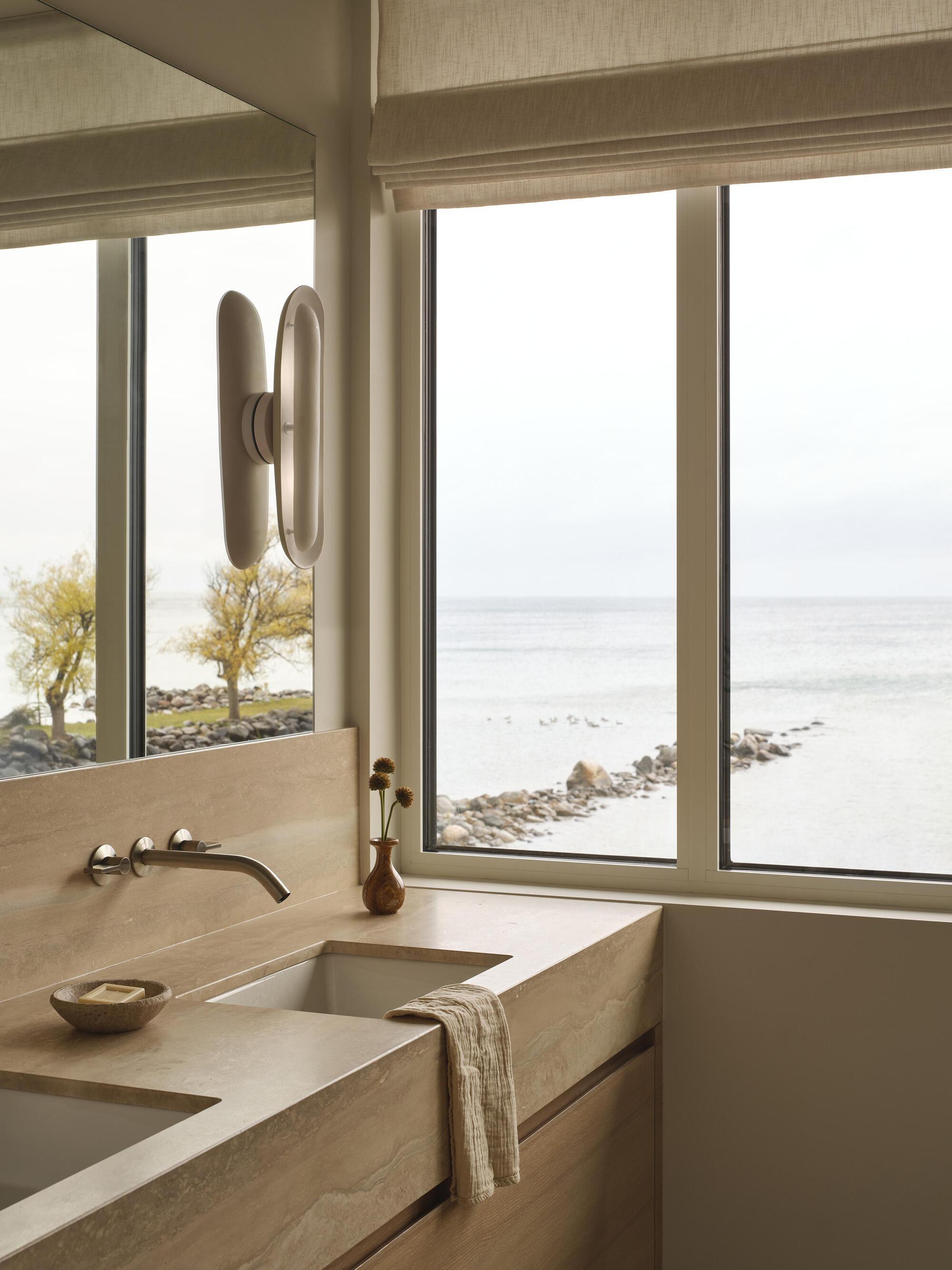 Bathroom vanity in pale stone with wall-mounted faucet facing windows overlooking the waterfront