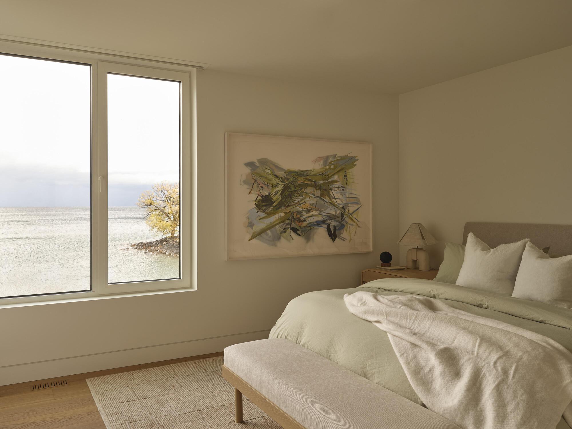 Bedroom with corner window framing a lone tree and distant water under overcast sky