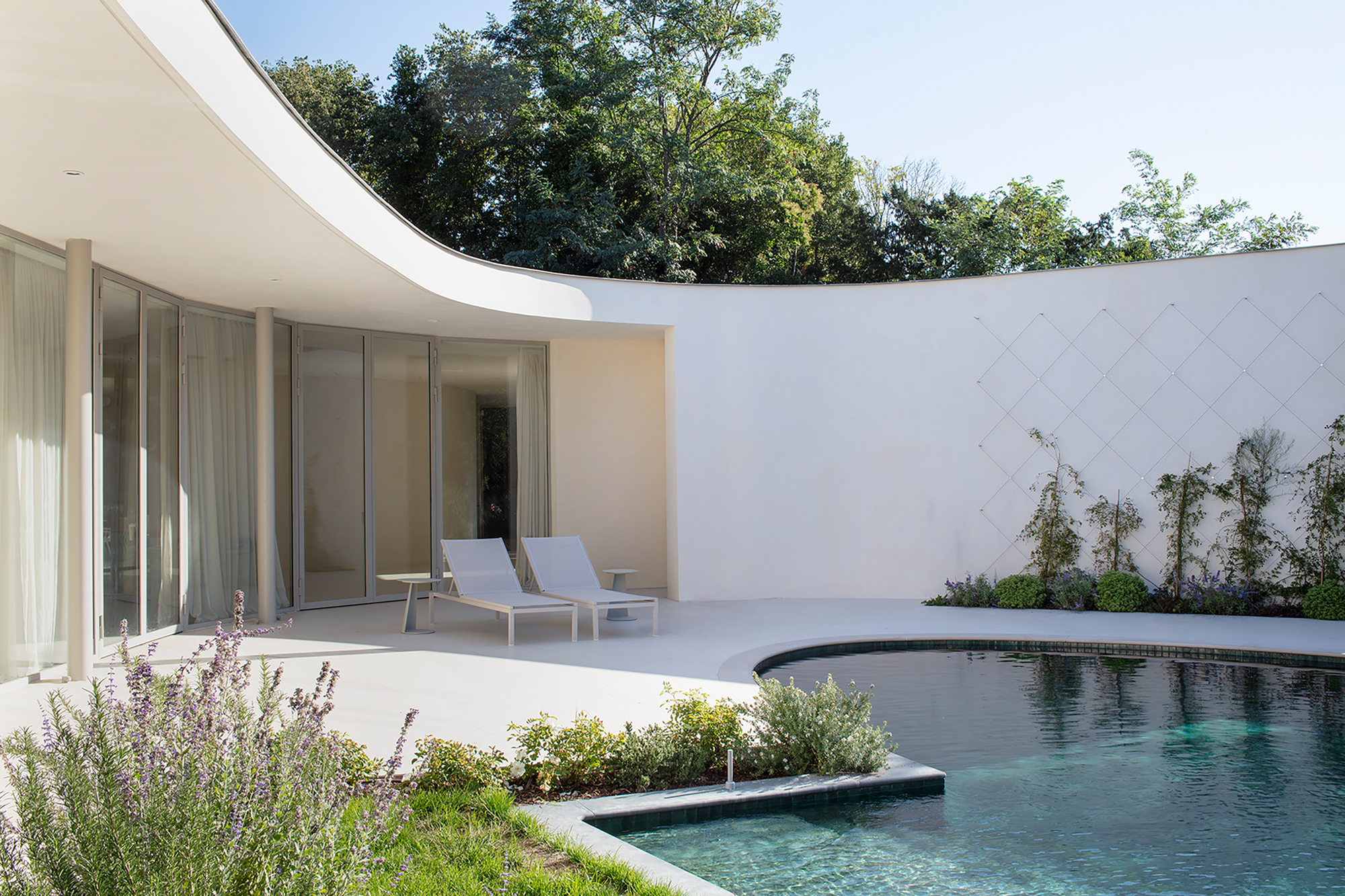 Pool terrace with glass pavilion, curved overhang, and planted beds along white tiled wall