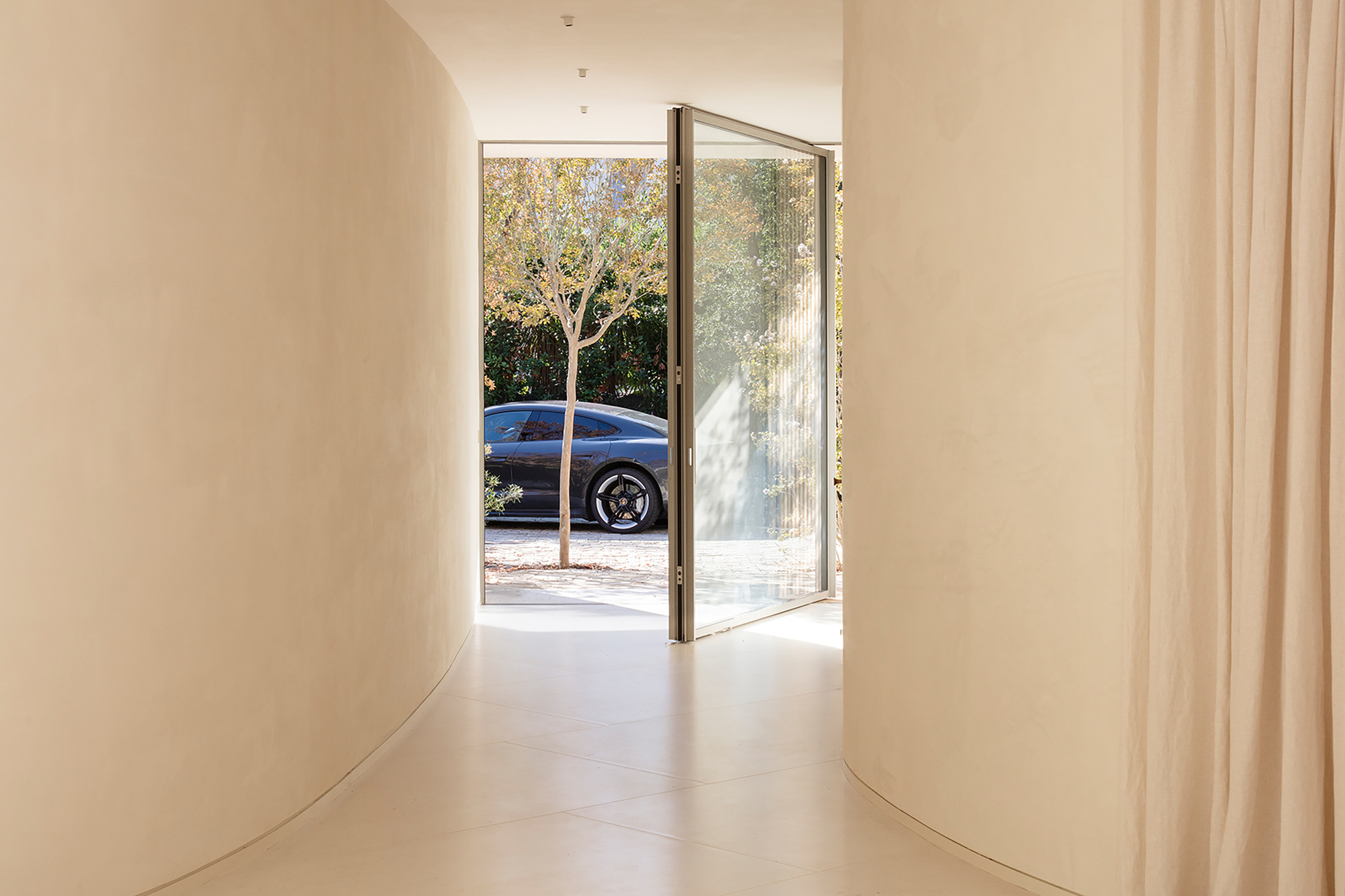 Curved hallway entrance with glass pivot door framing a blue car and tree outside