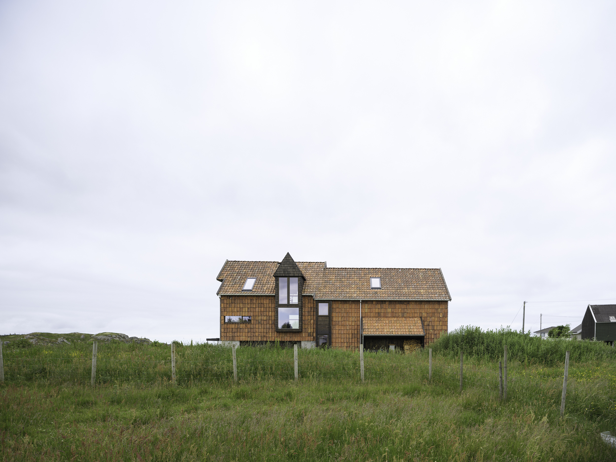 Timber-clad gabled house on a hillside with fenced pasture under overcast sky