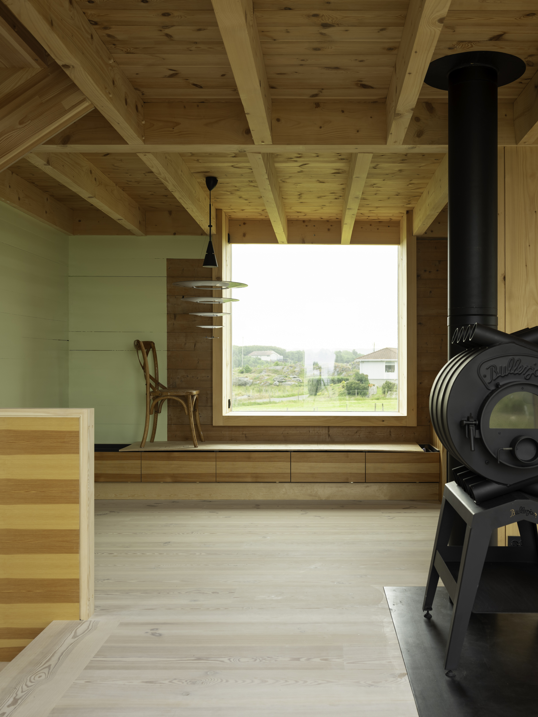 Interior with exposed timber ceiling beams and a black wood stove beside a window seat overlooking green fields