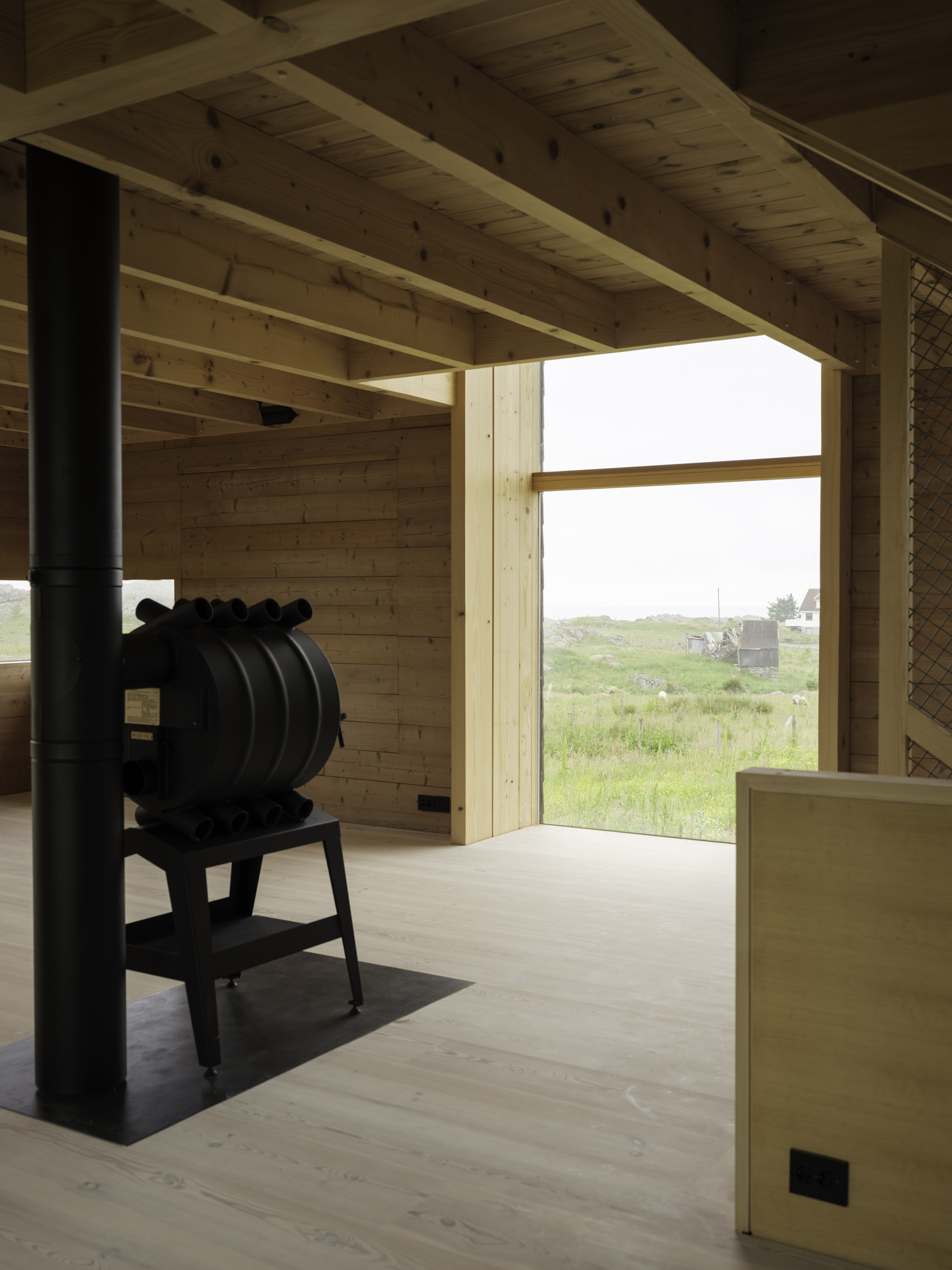 Black cast iron stove on a metal hearth beneath a timber beam ceiling with landscape view beyond