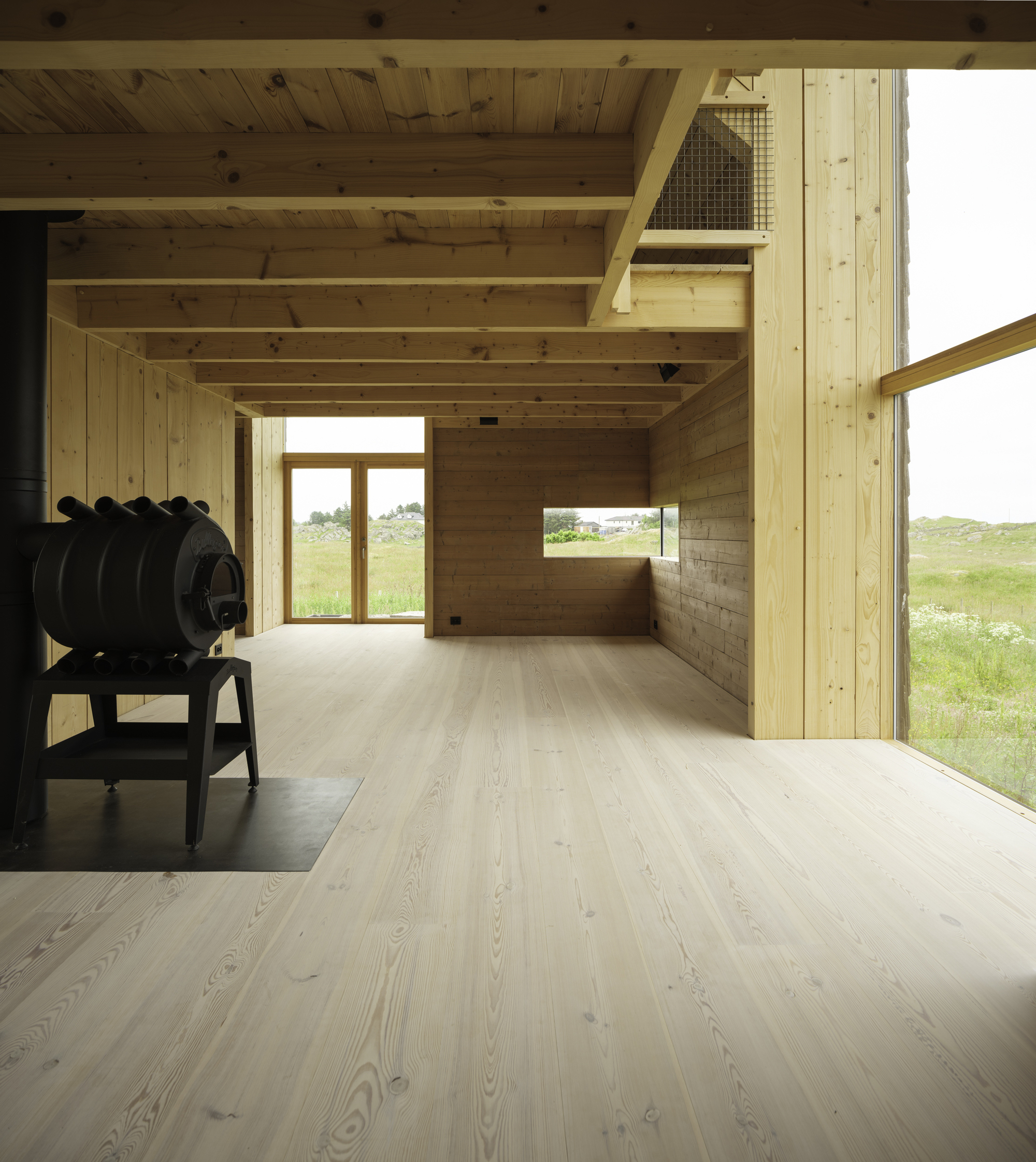Interior view of timber room with exposed beams and a black wood stove overlooking grassy landscape
