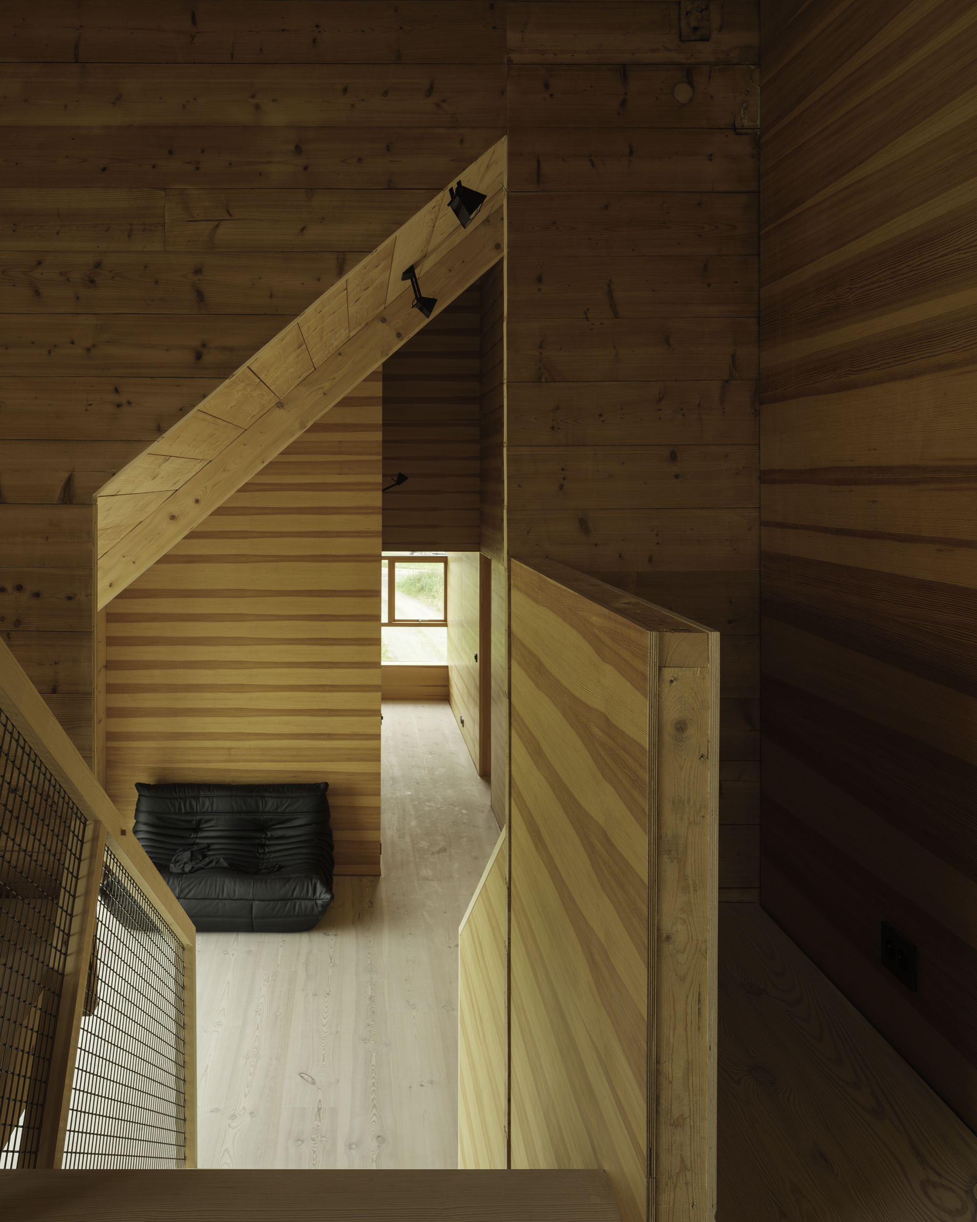 View down a double-height timber-lined corridor with diagonal wood slat stair rising alongside a black leather bench