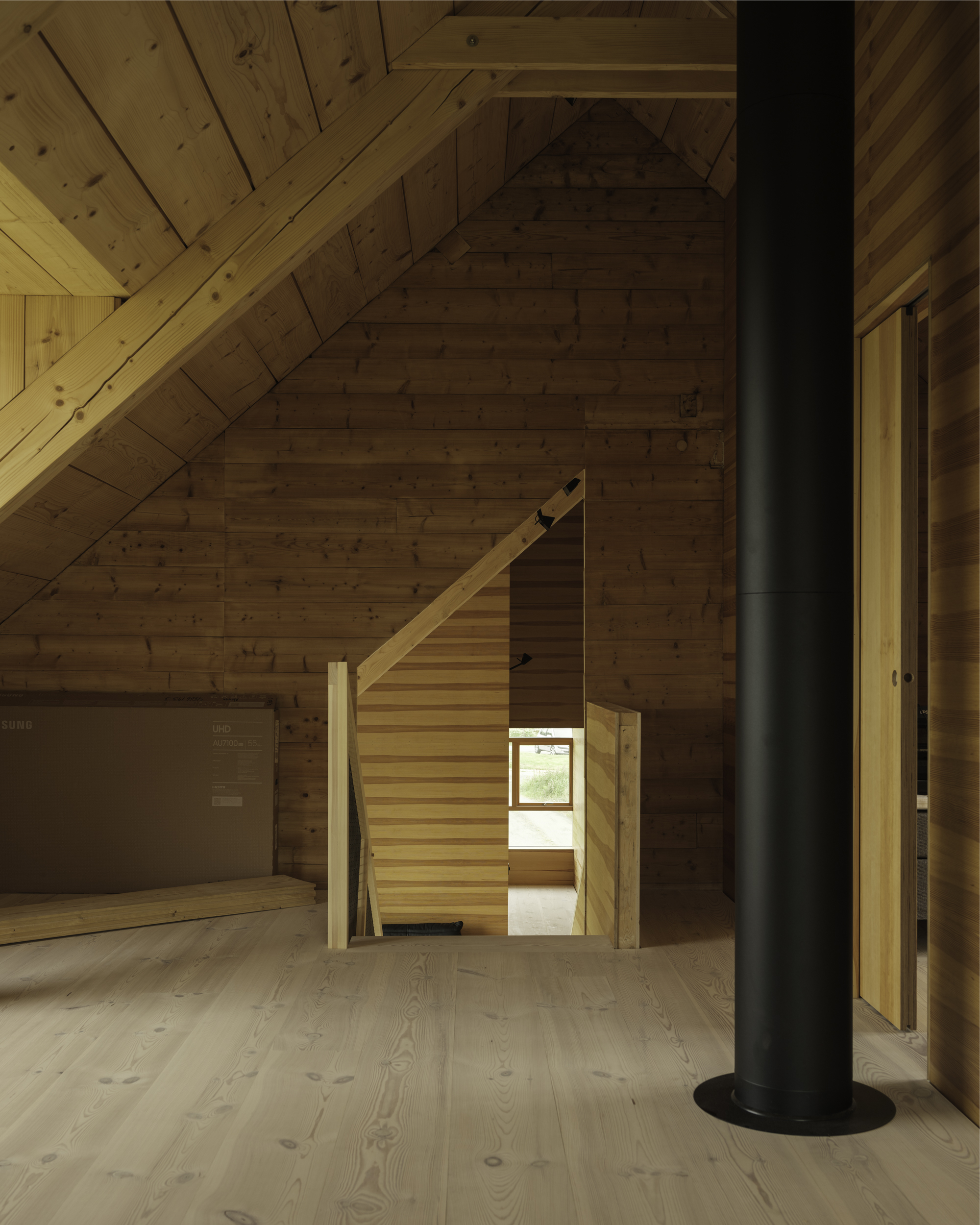 Timber interior with exposed diagonal ceiling planks, freestanding black flue pipe, and open stairwell beyond doorway