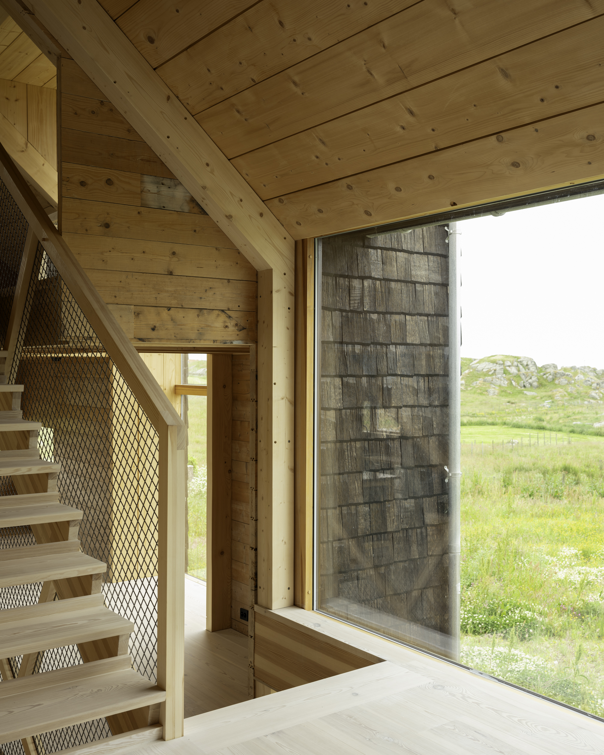 Open stair with mesh guardrail next to full-height glazing overlooking green meadow under overcast sky