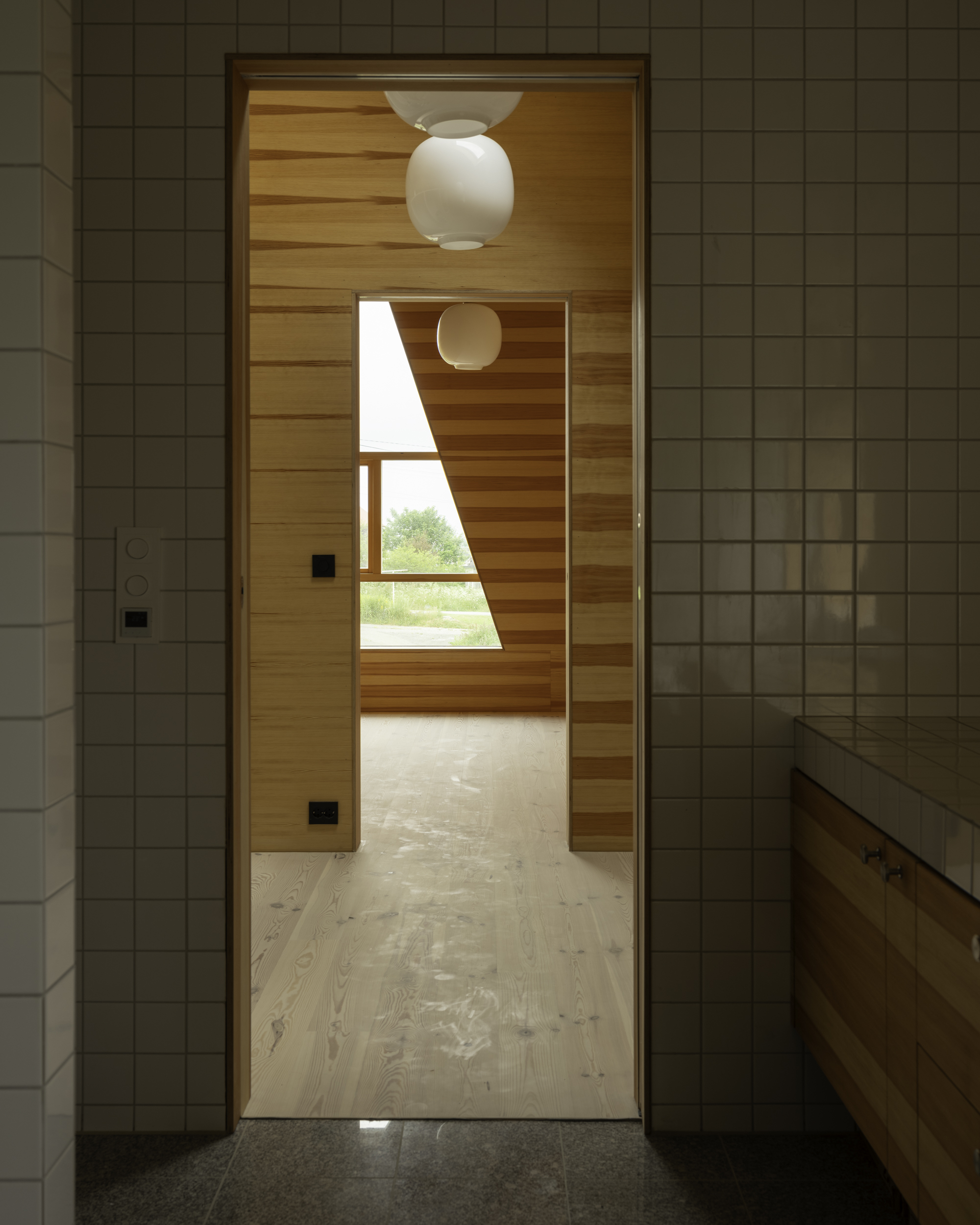 Doorway from white tiled bathroom into timber hallway with spherical pendant lamp and angled window framing greenery