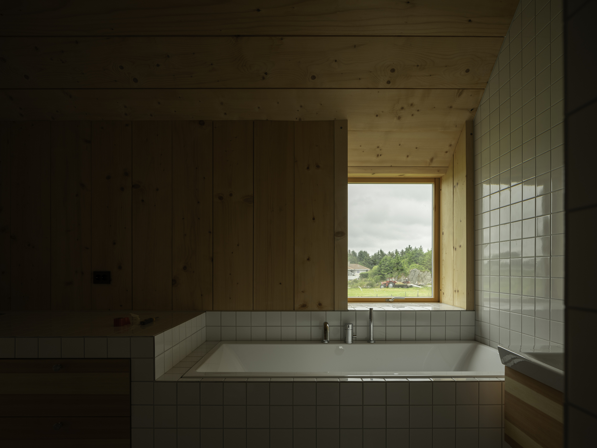 Recessed white-tiled bathtub beneath a timber-framed window overlooking distant forest and shoreline