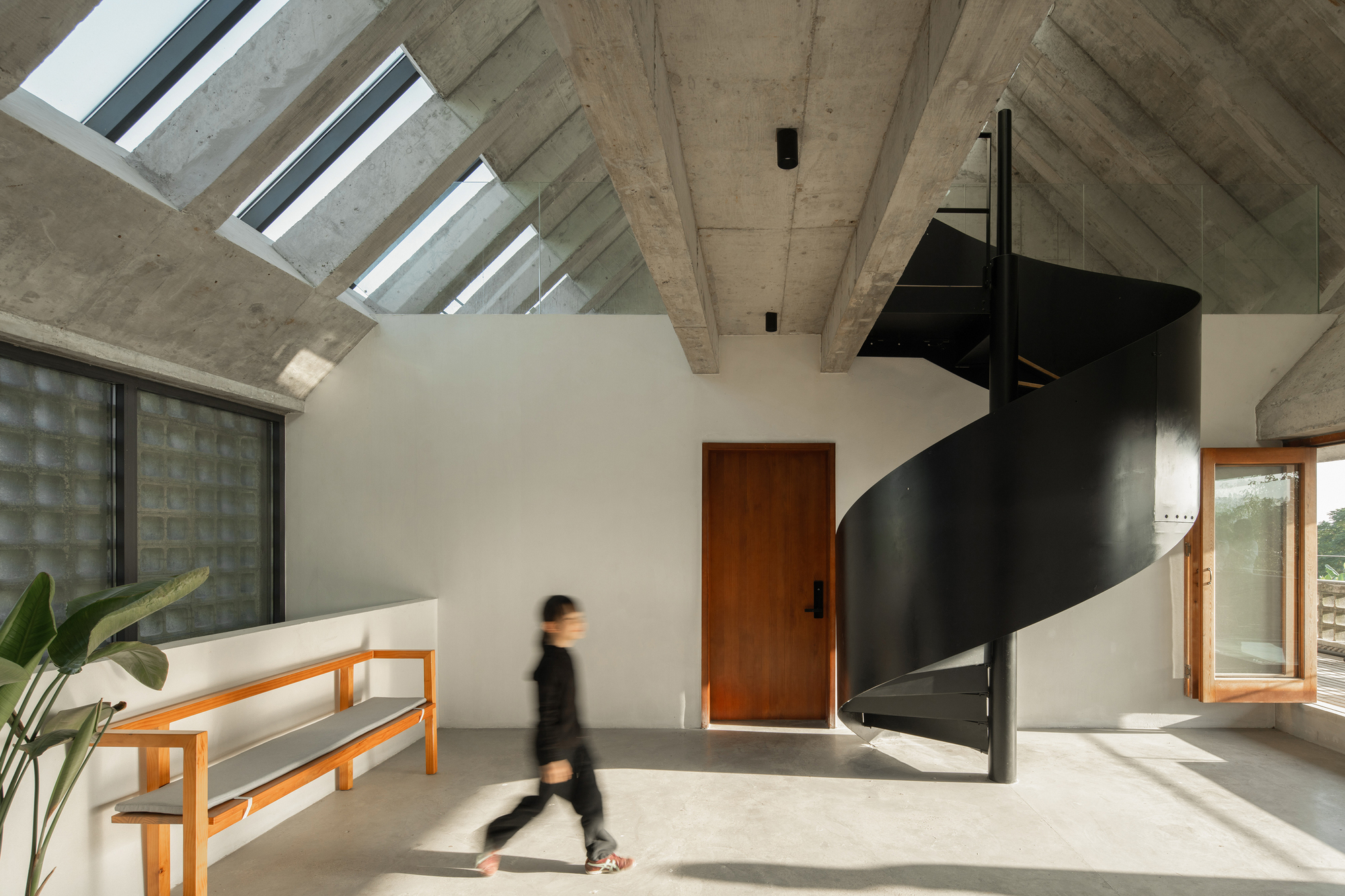 Double-height interior with black spiral staircase and skylight beams casting shadows as a figure walks through