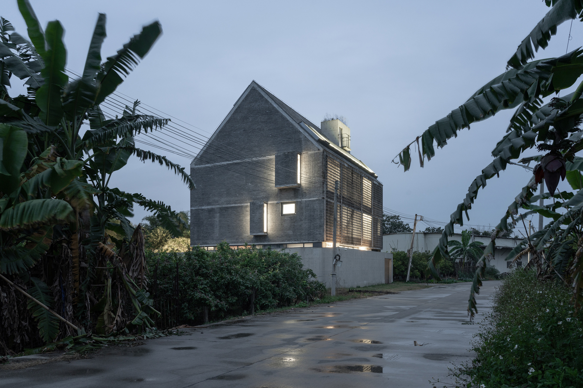 Gable-roofed concrete house with illuminated slit windows viewed from street flanked by banana plants