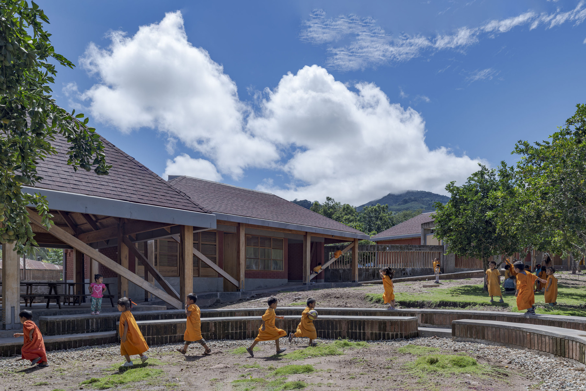 Courtyard with low-slung pitched roof buildings and stepped timber retaining walls as children walk along the pathways