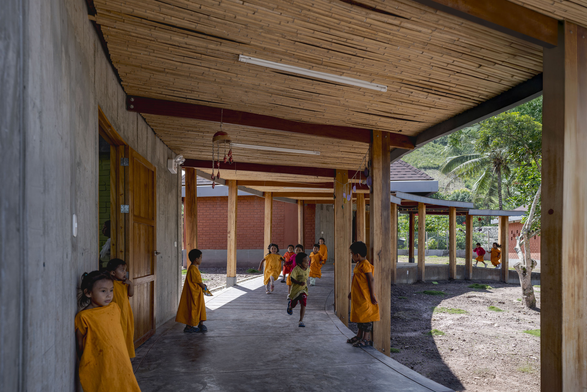 Covered walkway with timber columns and exposed ceiling beams, children in yellow uniforms running through dappled sunlight