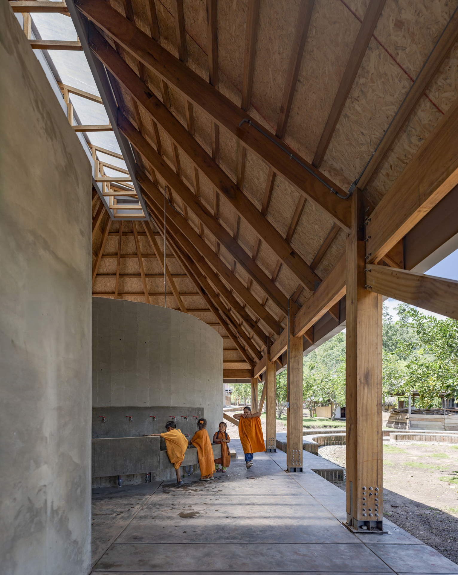 Open-air corridor with sloped timber ceiling panels, concrete columns, and children walking beneath the eaves