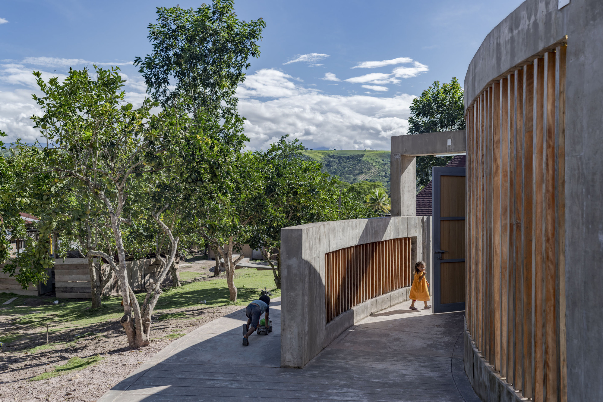 Elevated concrete walkway with vertical timber screens, children walking beside trees under a cloudy sky