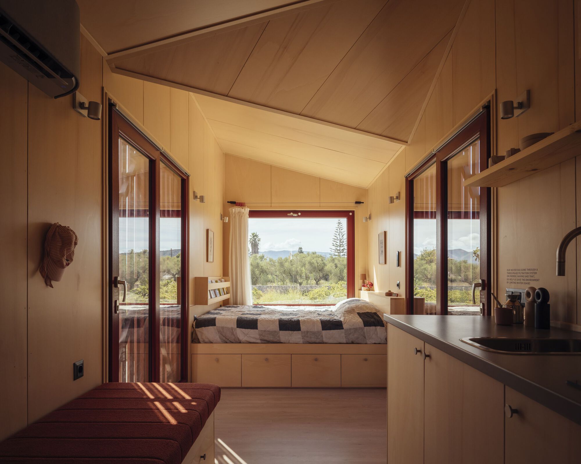 Interior view through plywood-lined space with glazed doors opening to a bed nook and landscape beyond