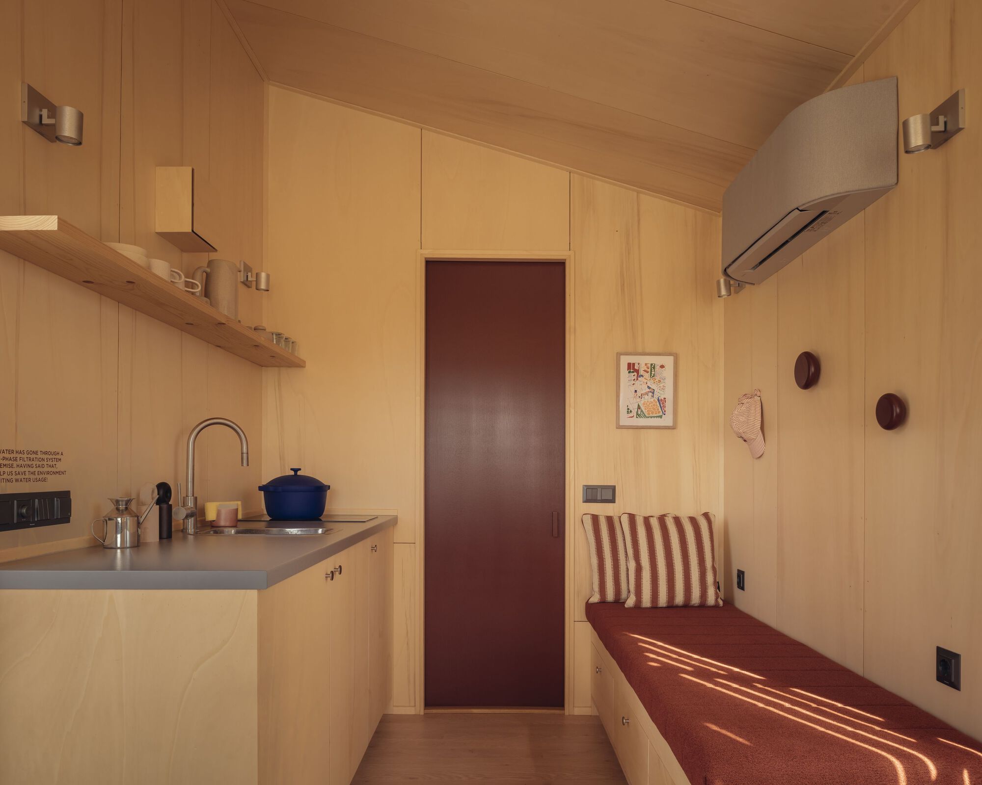 Compact kitchenette with birch plywood walls, dark red door and built-in upholstered bench in afternoon light