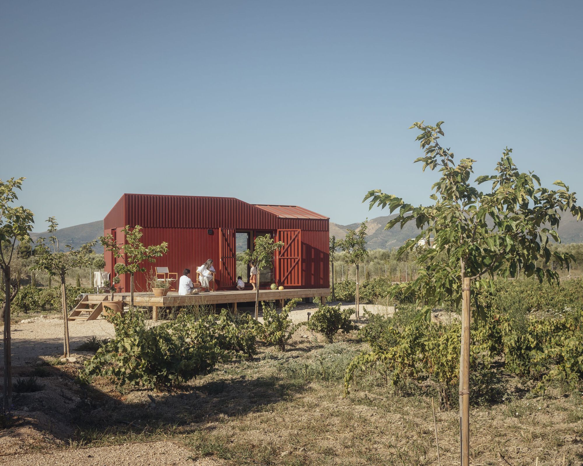 Timber deck with seated figures in front of the red metal structure and young orchard