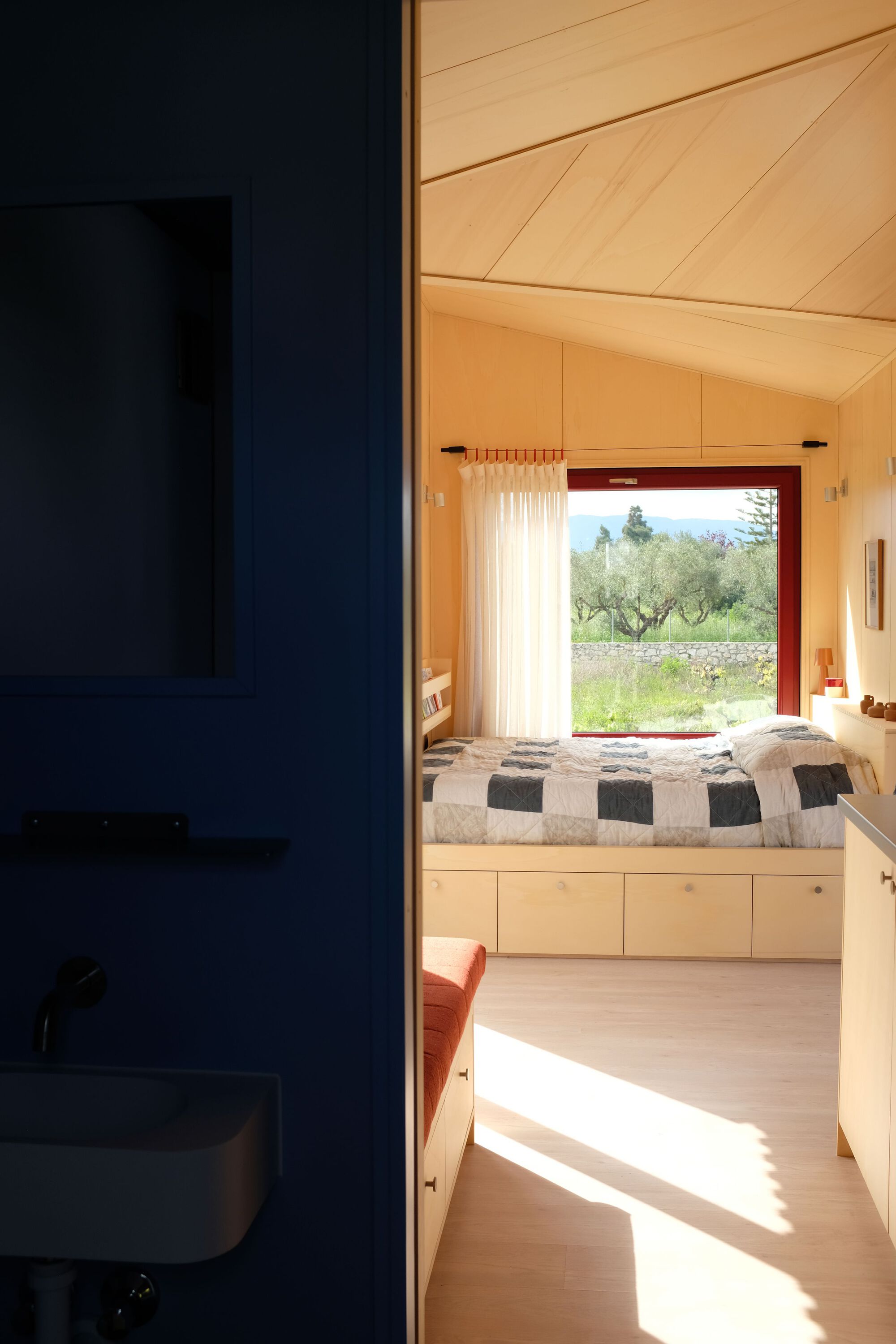View from dark blue bathroom into bedroom with sloped plywood ceiling and window opening to olive grove