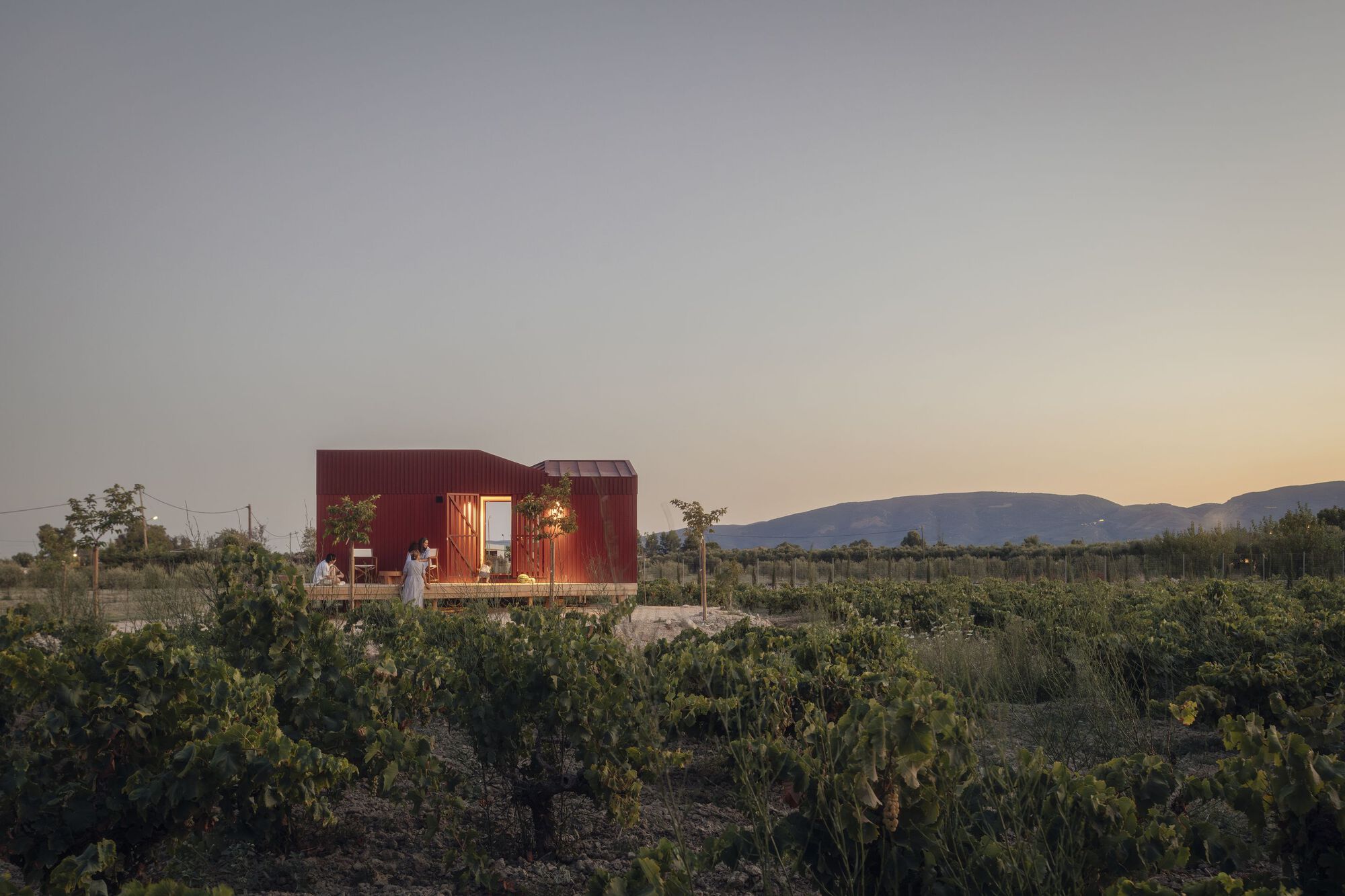 Red corrugated metal pavilion with open side opening set in a scrubland field at dusk