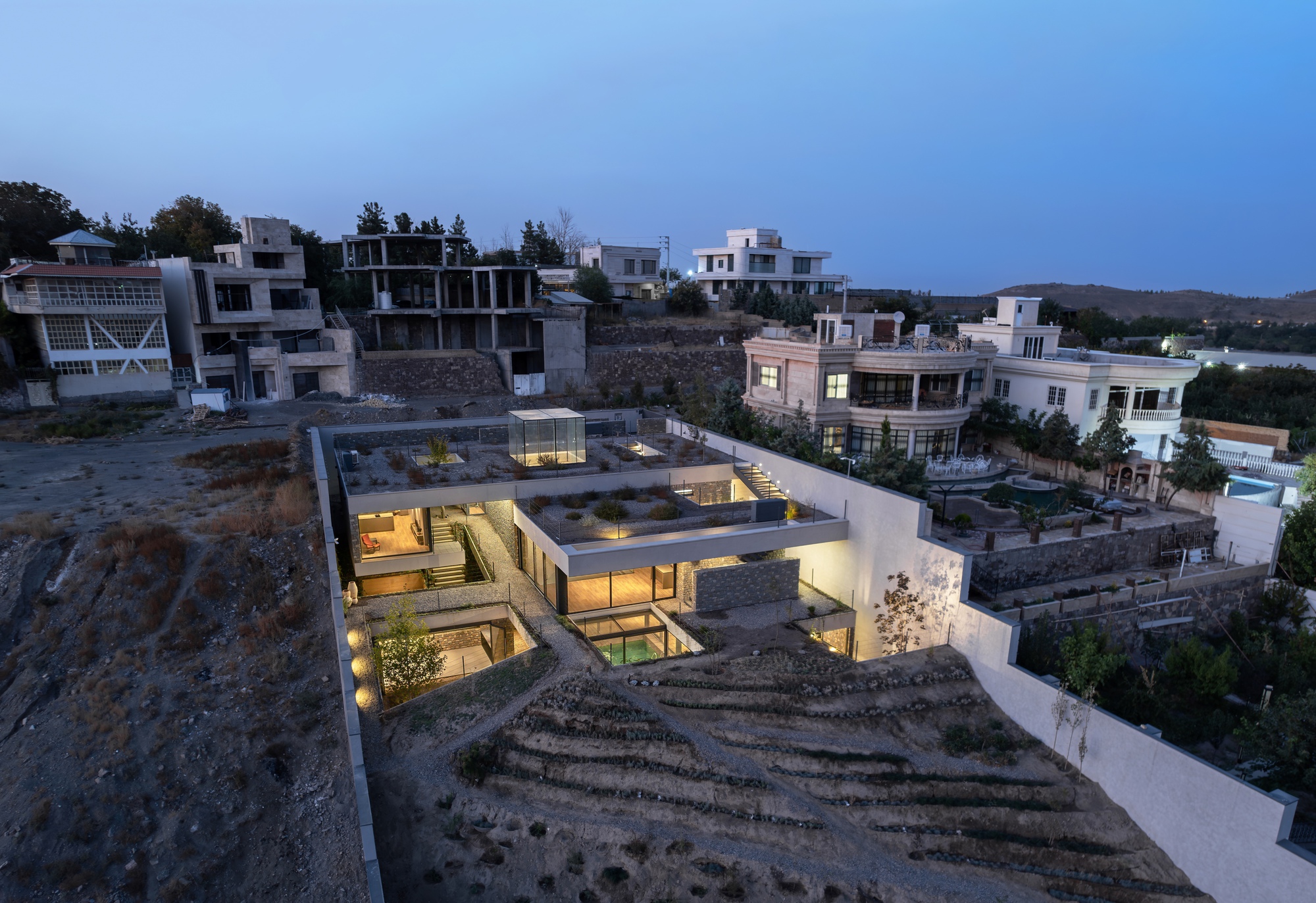 Aerial view of the low-slung residence with illuminated interior courtyards on a terraced hillside at dusk