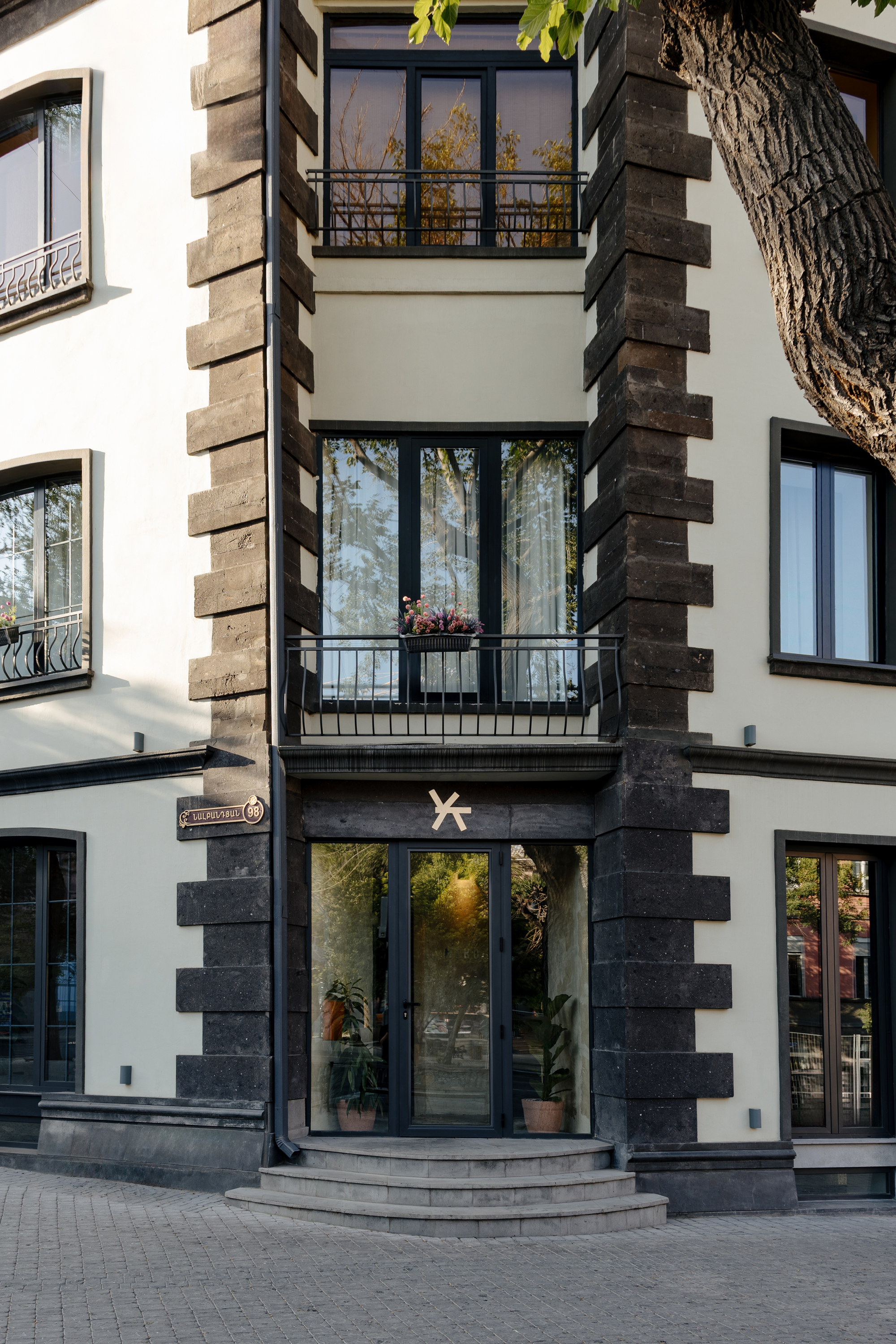 Street facade with white plaster and dark stone quoins framing balconies and glazed entry beneath a tree