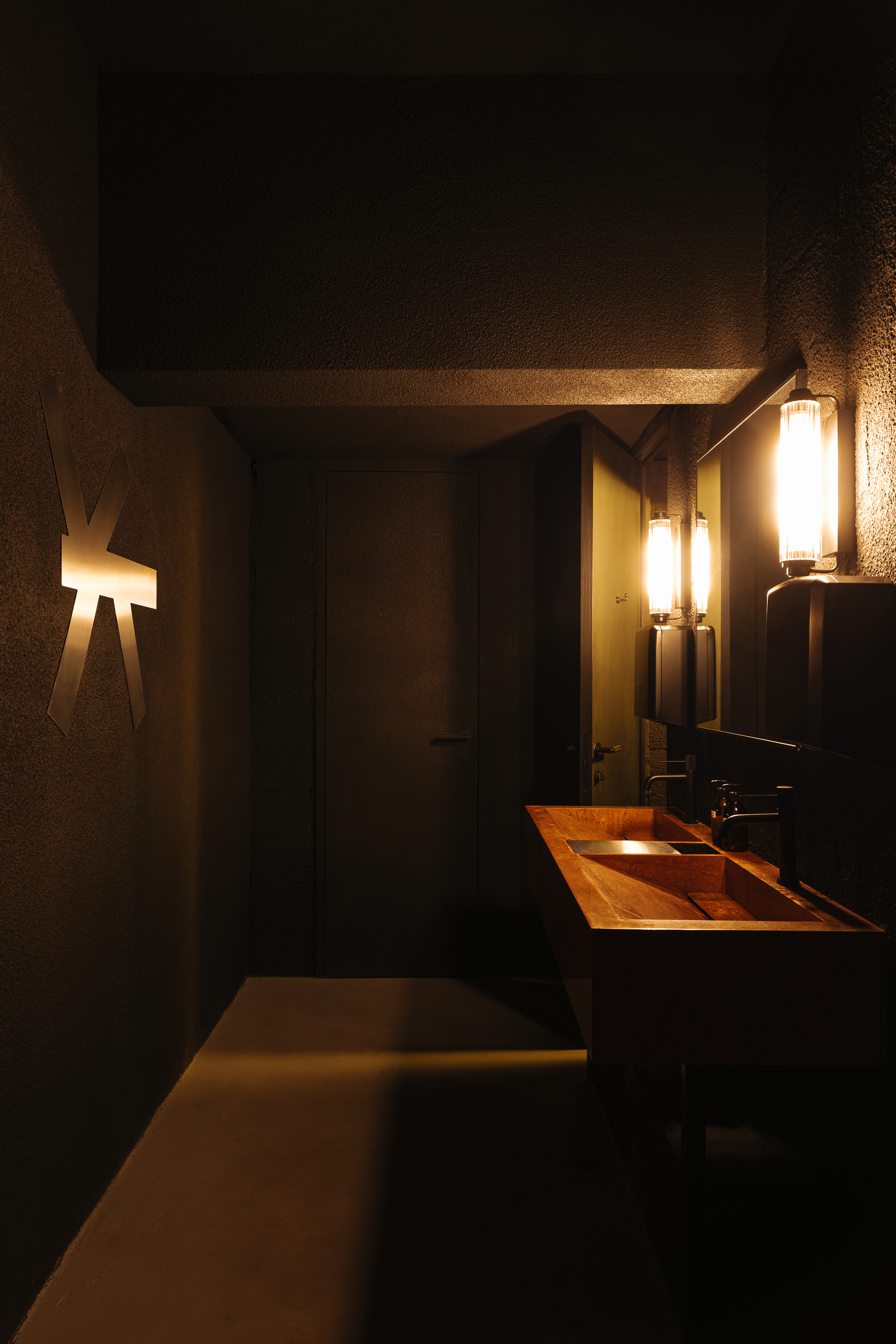 Restroom interior showing copper vanity, concrete ceiling and walls with wall sconces illuminating the space at night