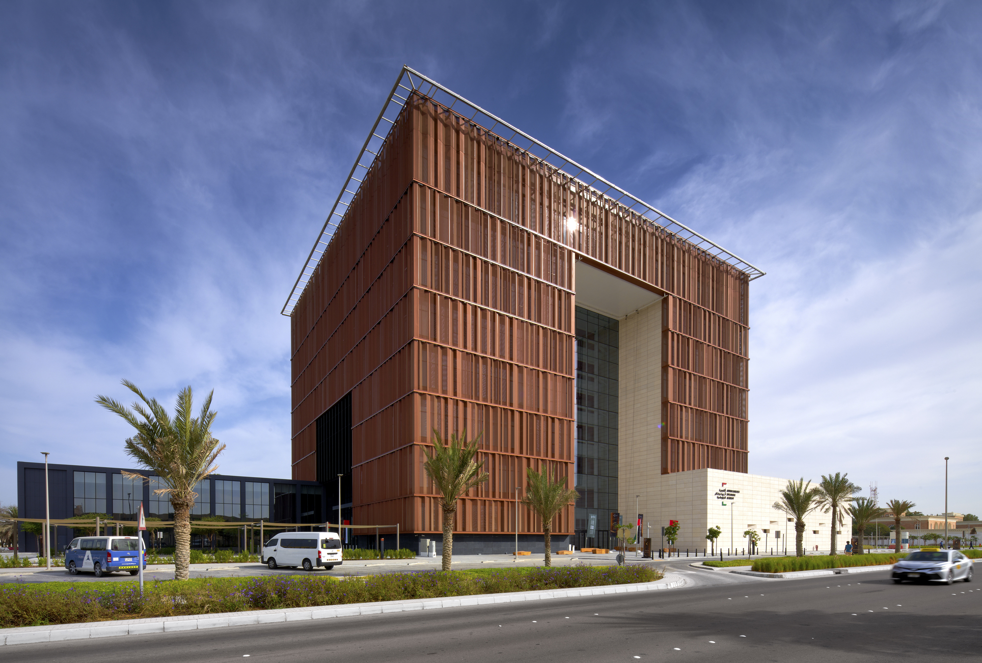 Cube-shaped volume clad in copper vertical louvers at a landscaped roundabout with palm trees