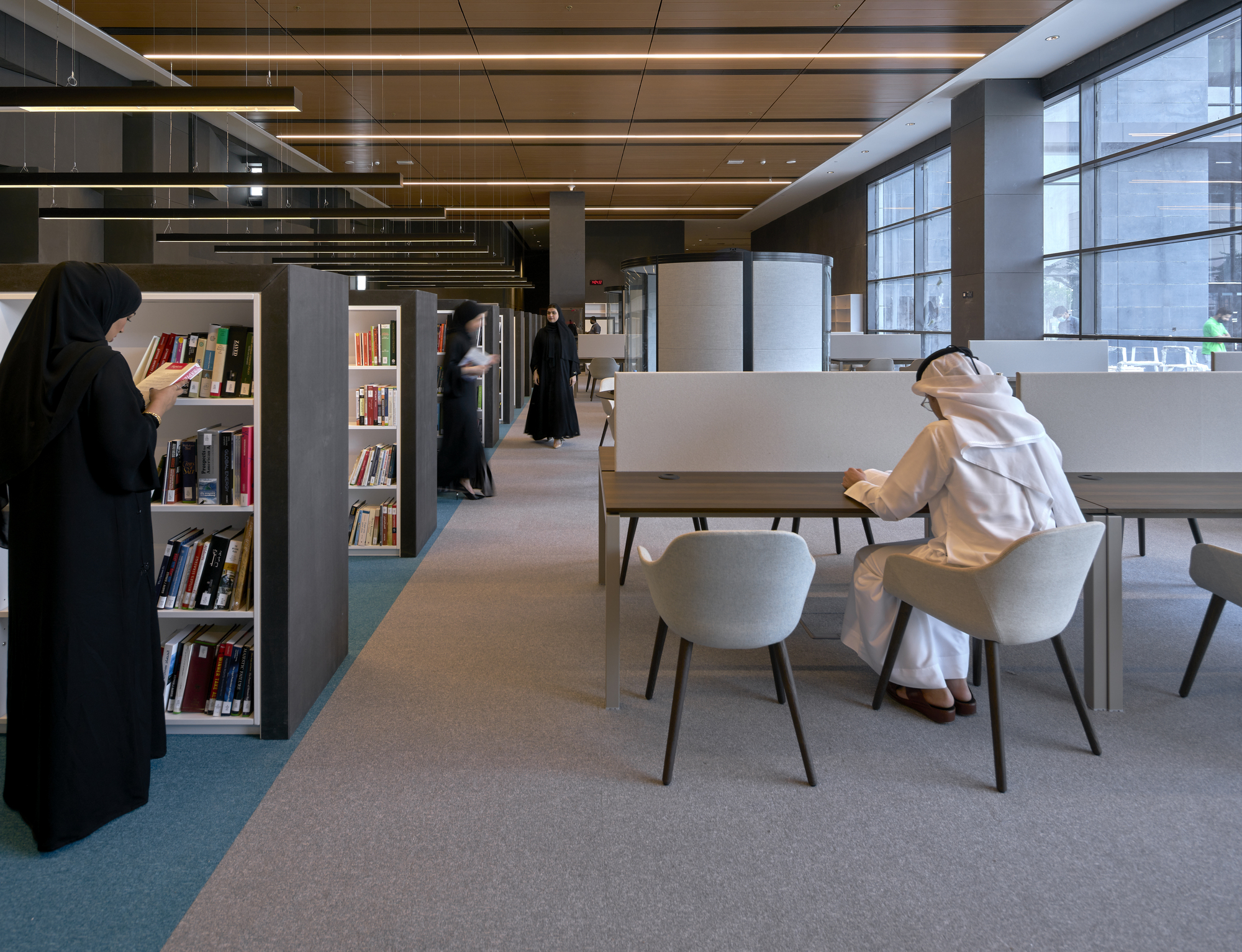 Library reading area with grey shelving and tables where visitors browse books beneath linear ceiling lights