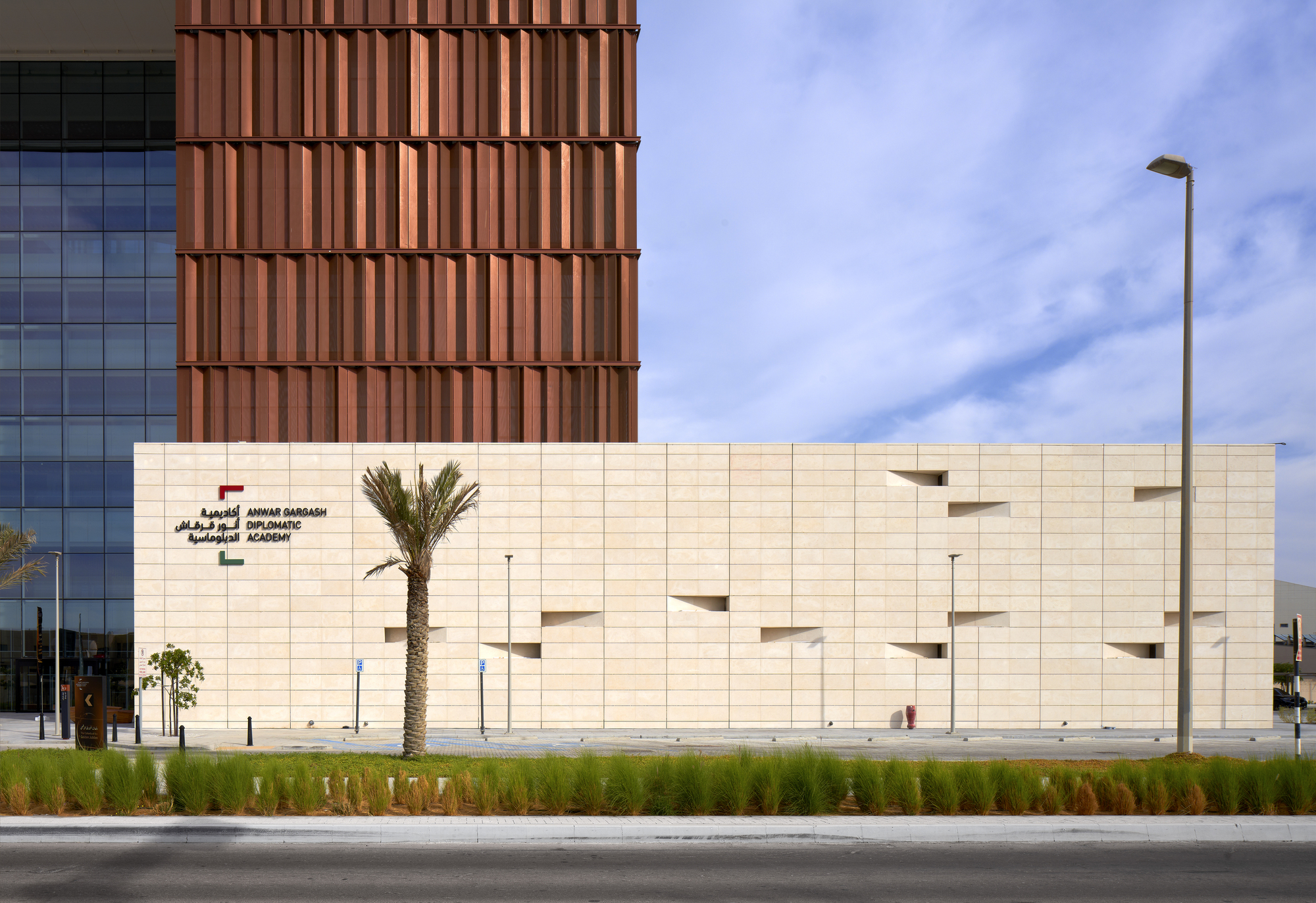Cream stone facade with recessed window openings beside a copper-clad tower and planted median under blue sky