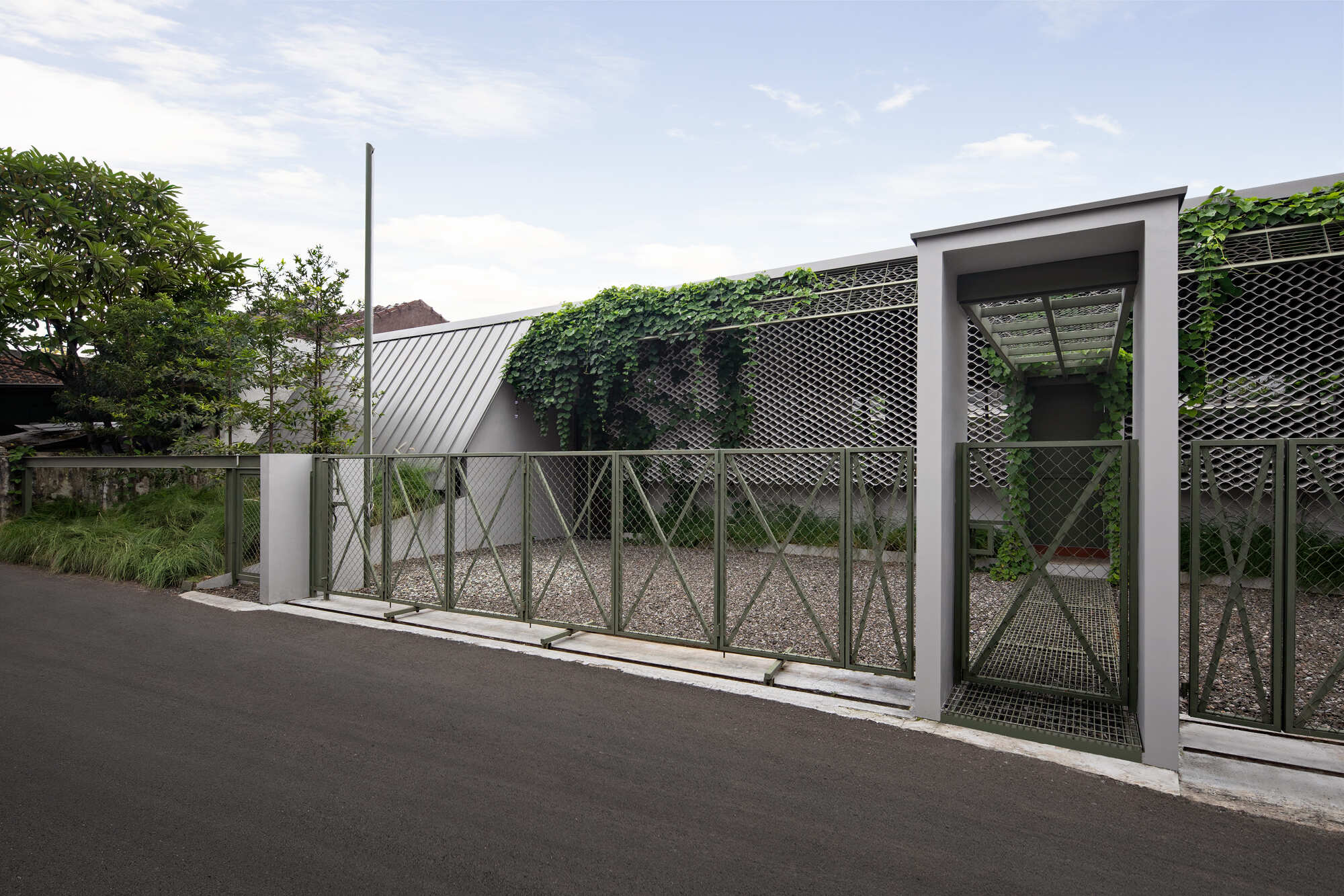 Street view of perforated metal fence with gate opening and climbing vines under a partly cloudy sky