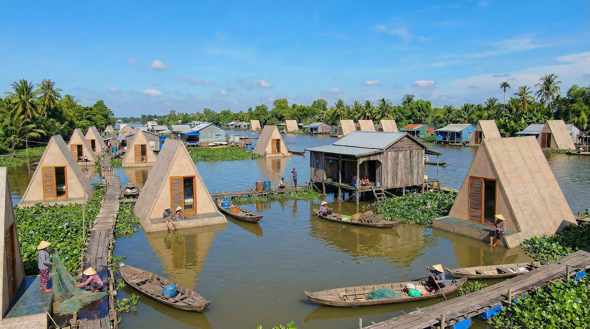 Triangular shelters with timber louvers along a wooden walkway in a flooded river settlement under clear skies