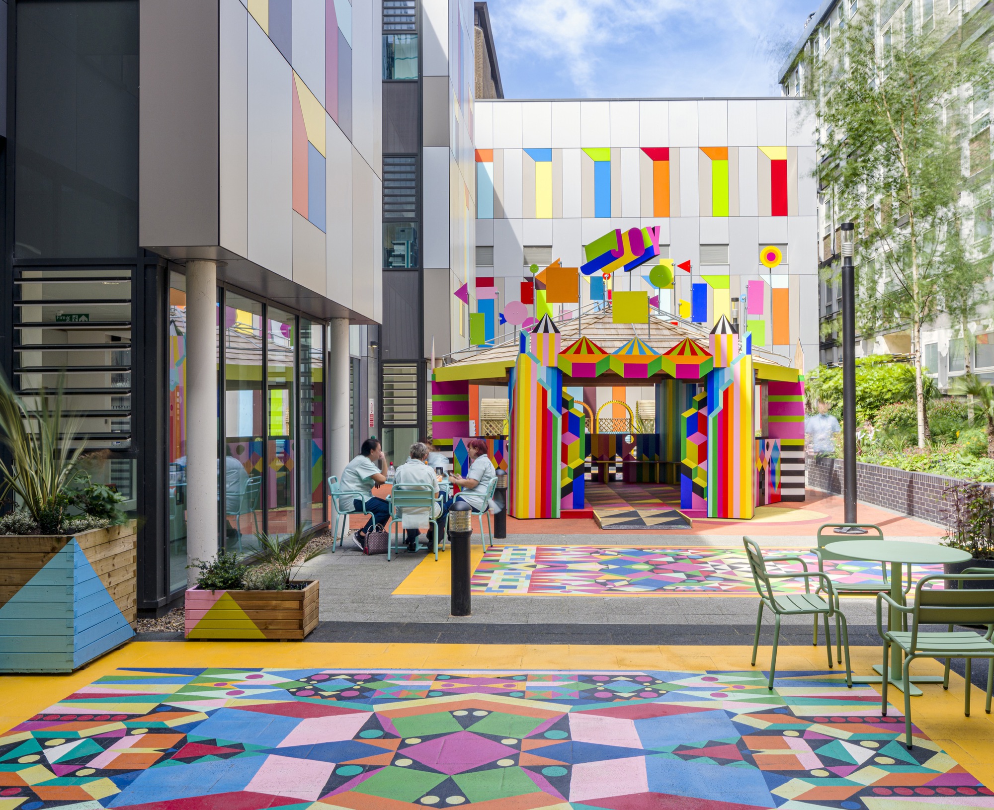 Courtyard with geometric paving and rainbow-striped pavilion entrance framed by residential buildings and diners at tables