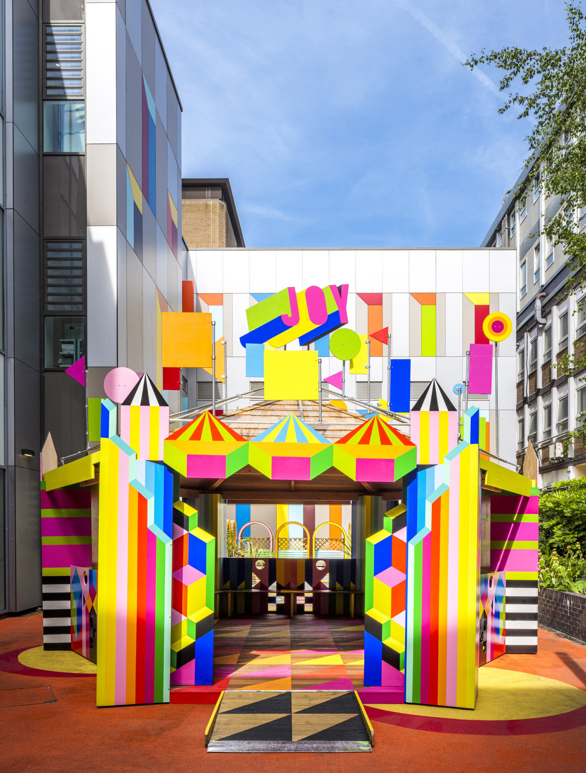 Frontal view of the pavilion showing timber roof, vertical striped columns and circular patterned paving under blue sky