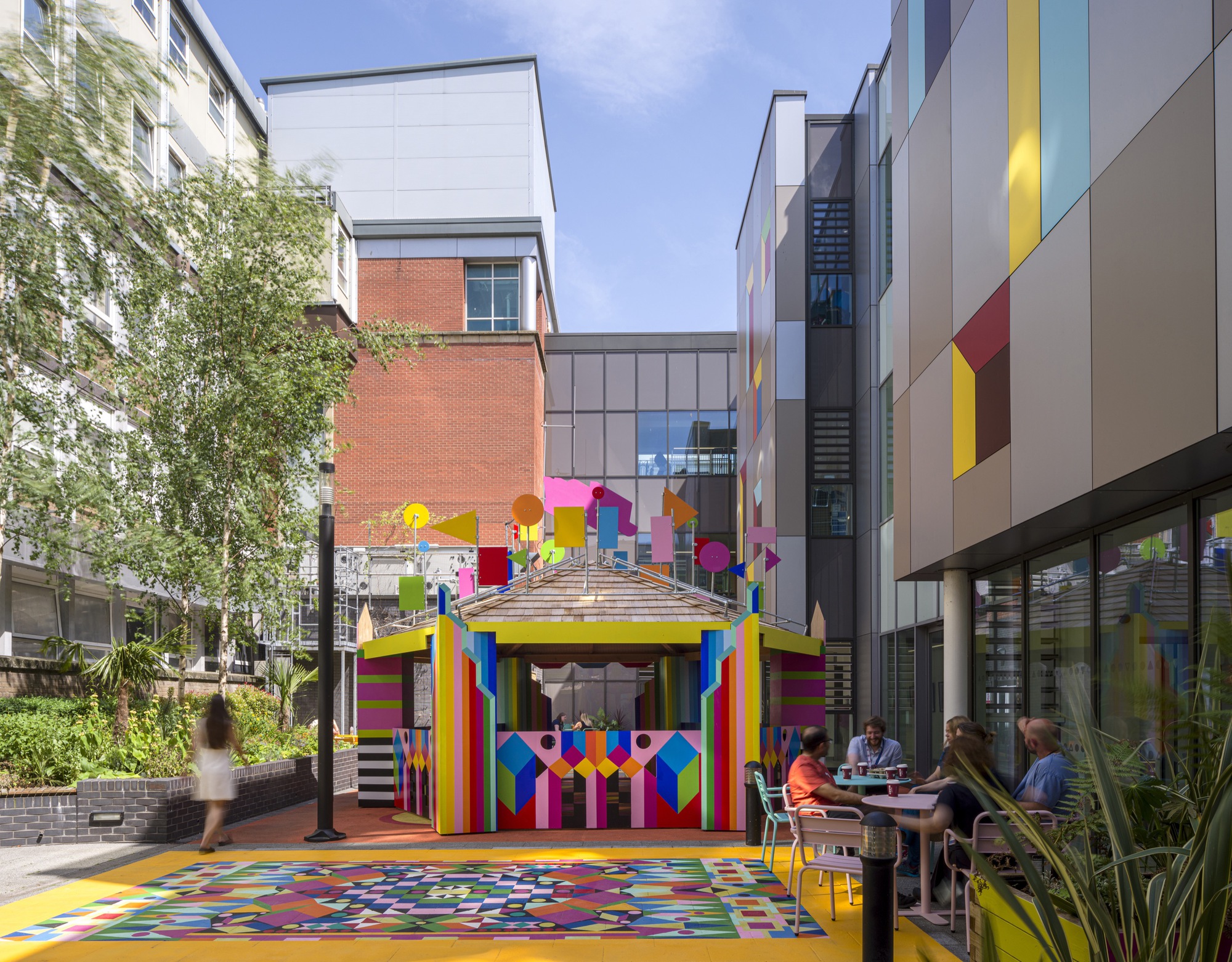 View across the courtyard showing the pavilion between residential blocks with pedestrians and seated groups on a sunny day